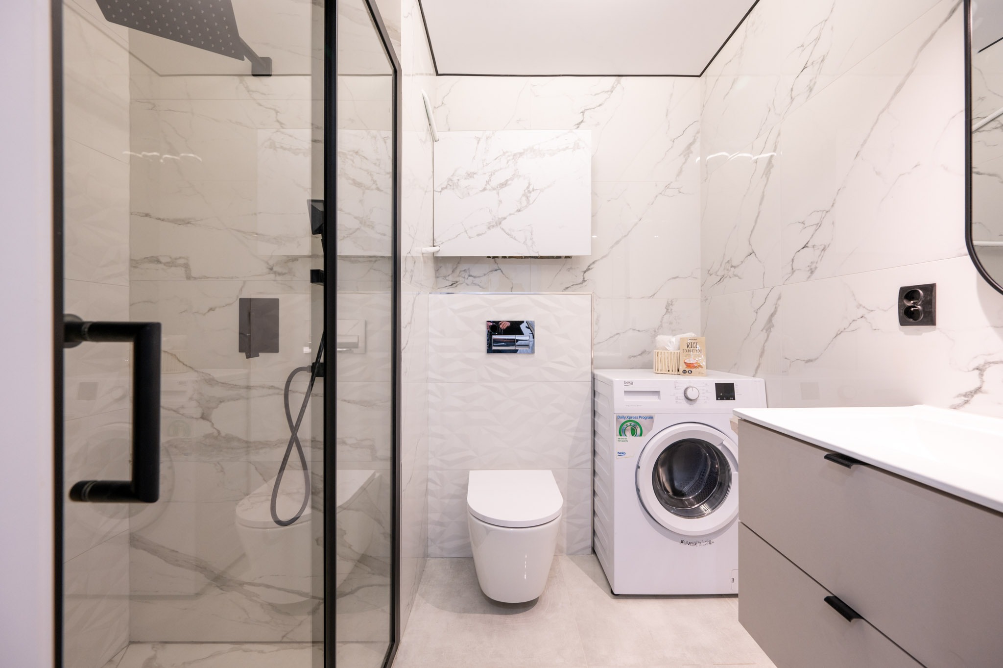 Nowoczesna łazienka z kabiną prysznicową typu walk-in, toaletą, pralką i szafką pod umywalką. Białe marmurowe płytki na ścianach i szare meble.