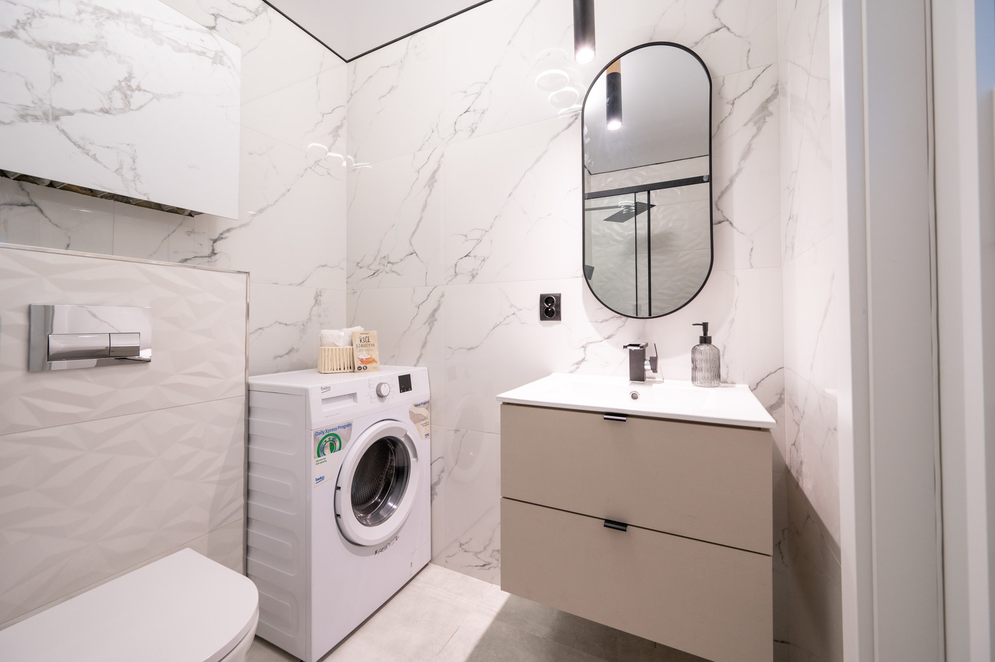 Nowoczesna łazienka z marmurowymi płytkami, beżową szafką pod umywalką, owalnym lustrem w czarnej ramie i pralką. Minimalistyczny design, stonowana kolorystyka.