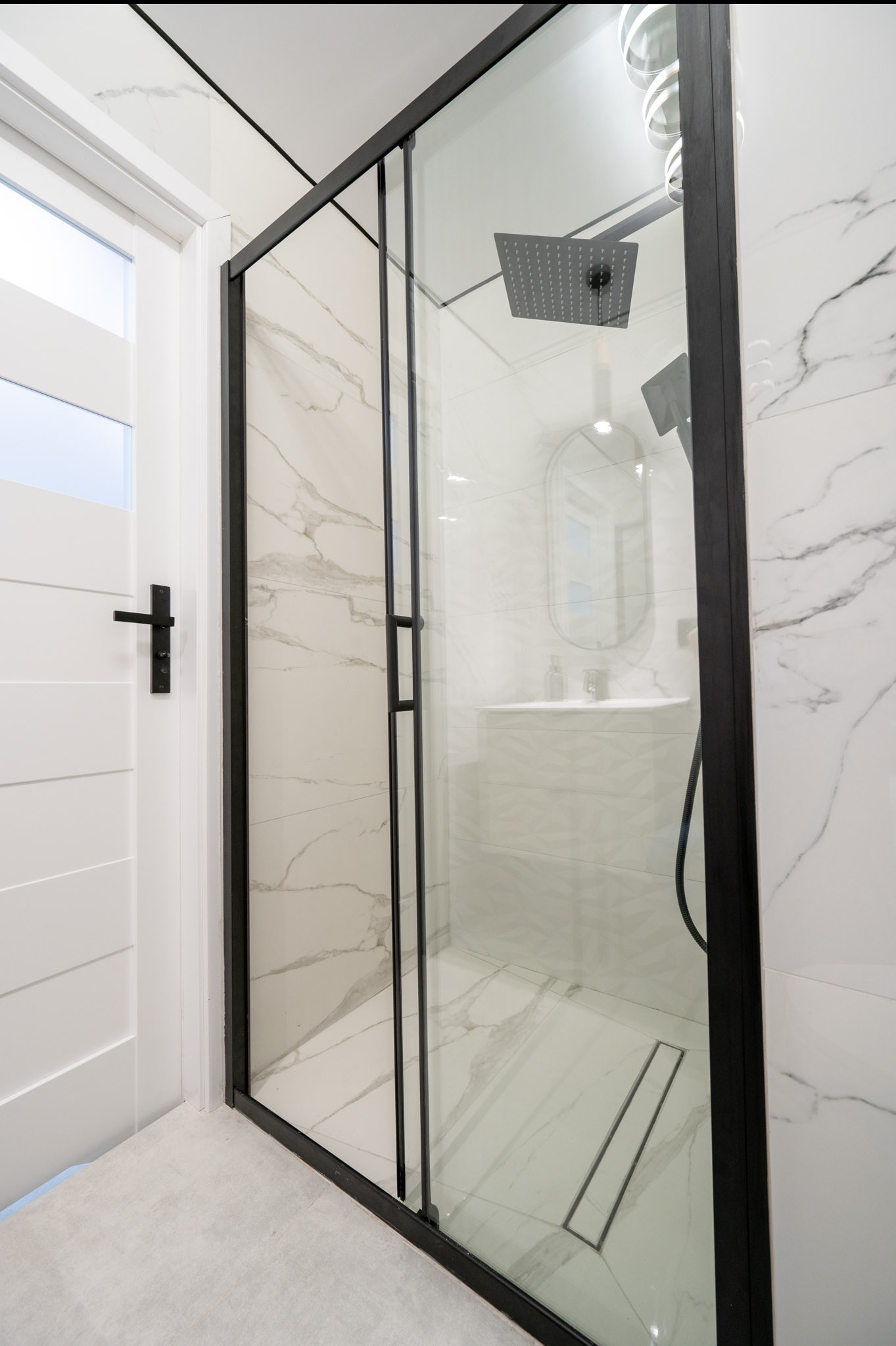 Nowoczesna kabina prysznicowa z czarnymi profilami i deszczownicą, białe płytki imitujące marmur, minimalistyczna armatura, widok z boku na drzwi do łazienki.