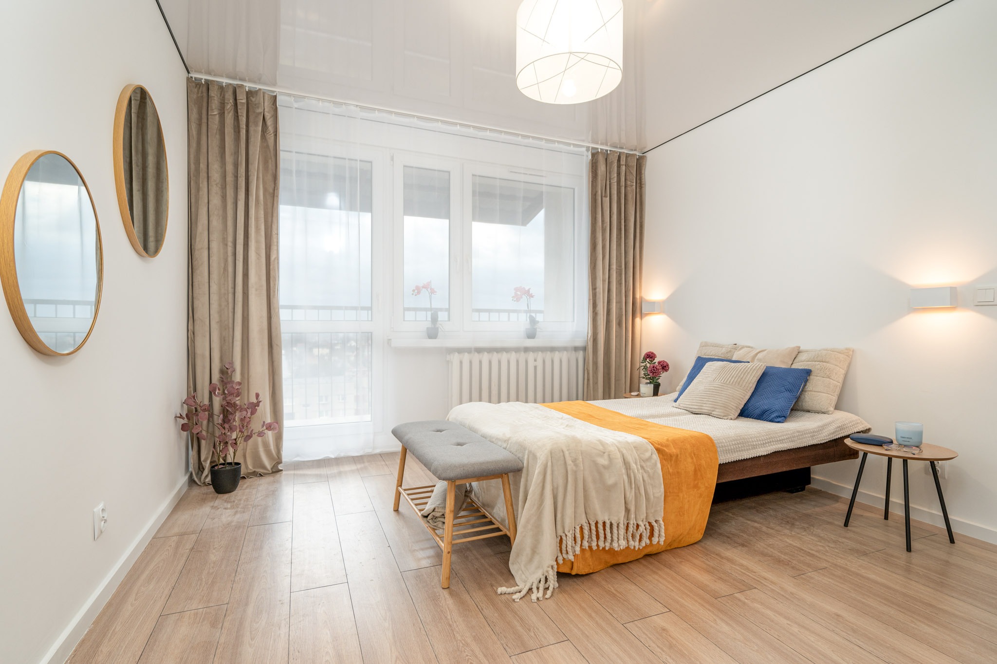 Jasna sypialnia z drewnianą podłogą i łóżkiem z żółtą narzutą. Dwa okrągłe lustra na ścianie. Minimalistyczny design, ciepłe oświetlenie i tekstylia tworzą przytulną atmosferę.