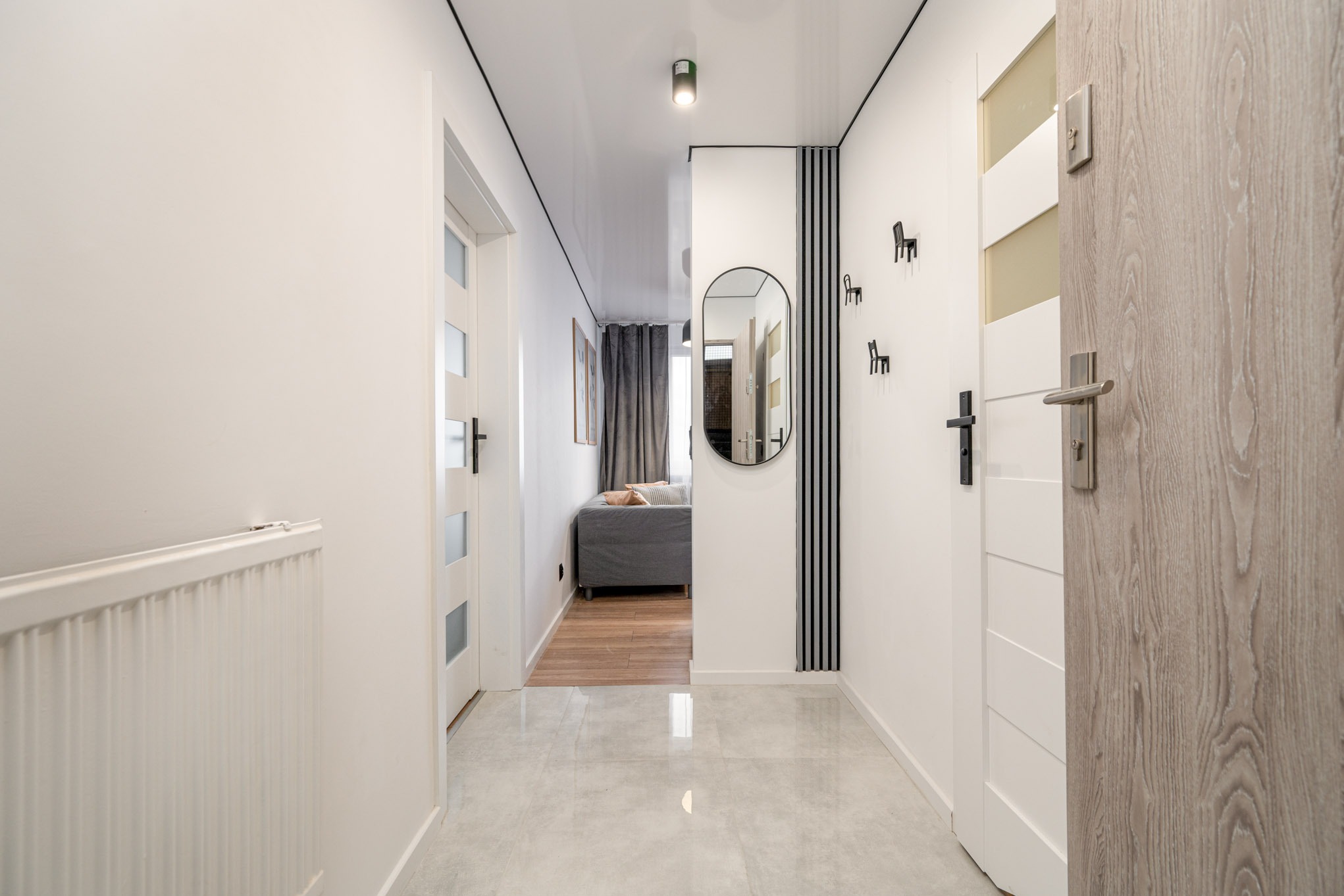 Nowoczesny przedpokój z błyszczącą podłogą, białymi ścianami, dekoracyjnym lustrem i drzwiami z przeszkleniami. Widoczny fragment salonu z szarą sofą.