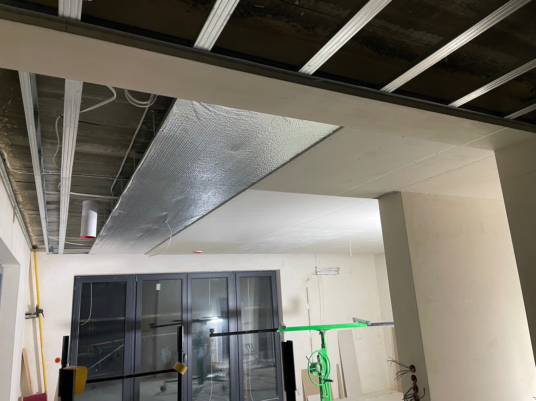 Sufit podwieszany z płyt karton-gips, częściowo z izolacją, widoczne profile i narzędzia montażowe. W tle okno balkonowe i instalacja elektryczna.