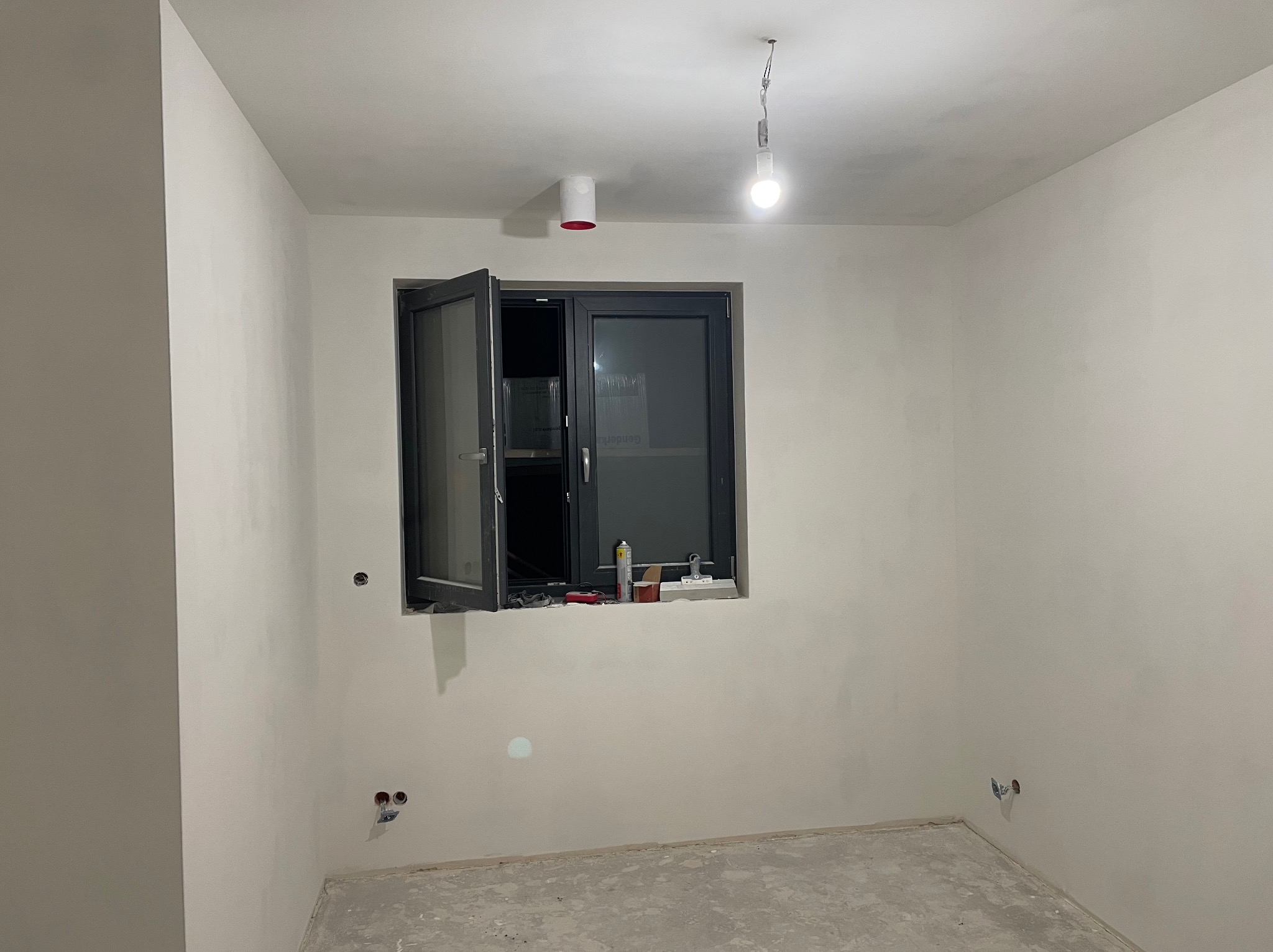 Pokój w trakcie prac wykończeniowych: szare okno z uchylonym skrzydłem, ściany po gładziowaniu, widoczne puszki elektryczne, surowa podłoga. Na parapecie narzędzia.