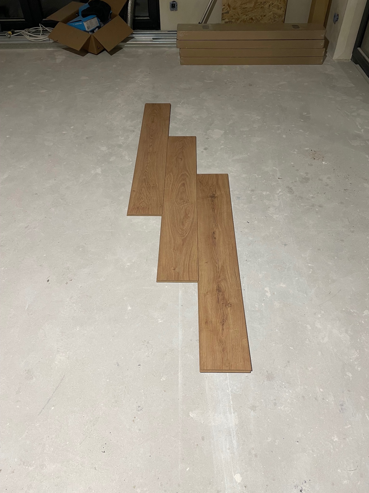 Układanie paneli podłogowych w surowym wnętrzu. Widoczne trzy panele z wyraźnym wzorem drewna na szarym, betonowym podłożu. W tle kartony z panelami i narzędzia.