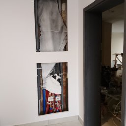 LD Oleksandr Laputin - Biała ściana z dwoma wnękami, w których widoczne są rury instalacyjne (woda, kanalizacja) i liczniki. Obok widoczne czarne futryny drzwiowe. W tle remont.