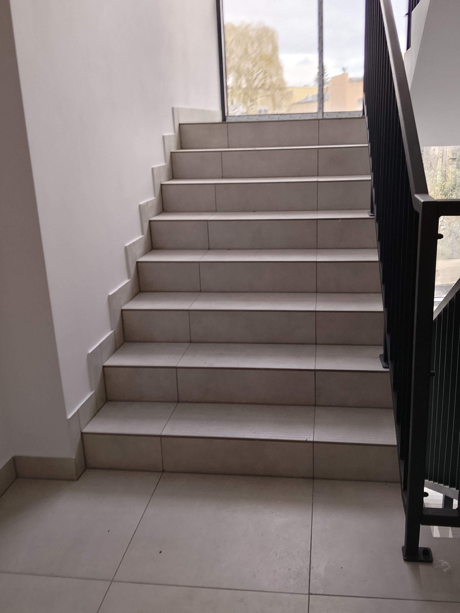 Wykończone schody z jasnymi płytkami i czarną balustradą w Jeleniej Górze. Widok z dołu, nowoczesny design, neutralne kolory, schludne wykończenie.