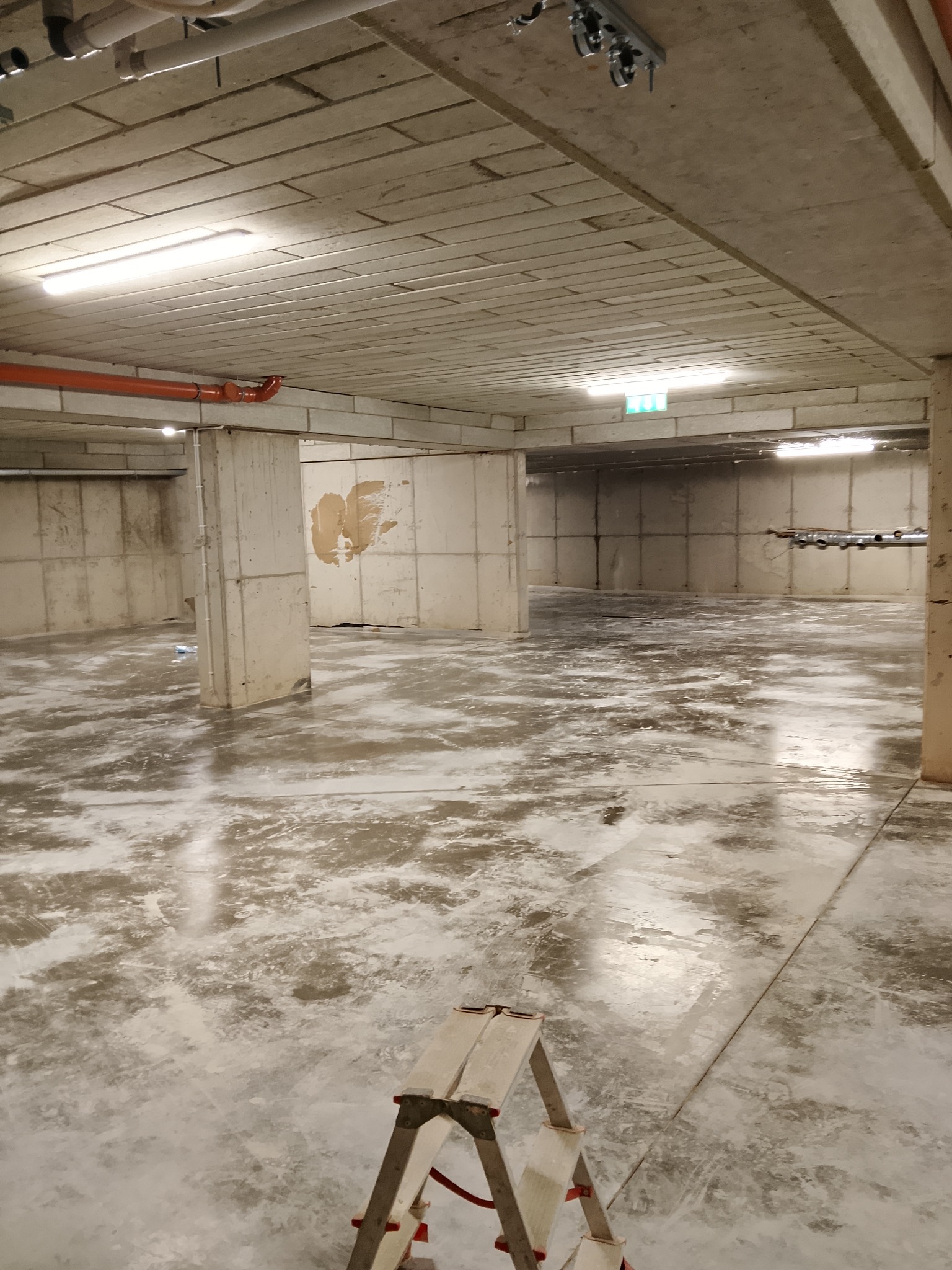 Surowe wnętrze z betonowymi ścianami i podłogą po wylewce, widoczne instalacje pod sufitem i drabina malarska w rogu, sugerujące prace wykończeniowe w toku.