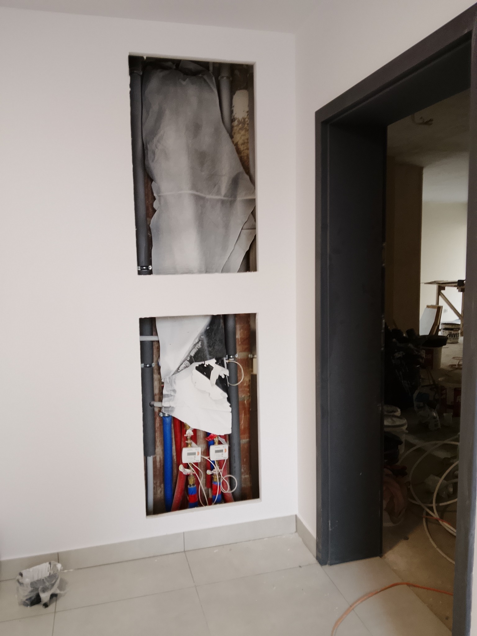 Biała ściana z dwoma wnękami, w których widoczne są rury instalacyjne (woda, kanalizacja) i liczniki. Obok widoczne czarne futryny drzwiowe. W tle remont.