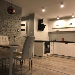 Krzysztof Dratwa - Nowoczesna kuchnia z białymi szafkami, czarnym blatem i okapem. Na pierwszym planie stół z krzesłami i dekoracyjna ściana z kamienia. Całość oświetlona punktowymi lampami.