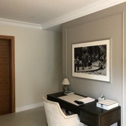 Krzysztof Dratwa - Pokój po malowaniu: beżowe ściany z białą sztukaterią, ciemne biurko z krzesłem w stylu glamour, na ścianie czarno-biała fotografia w ramie. Minimalistyczny design.