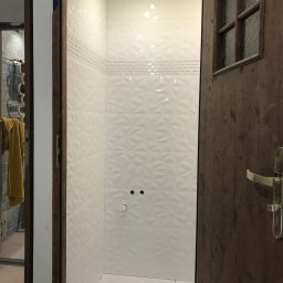 Krzysztof Dratwa - Wykończona kabina prysznicowa z białymi, trójwymiarowymi płytkami, widoczna przez otwarte, drewniane drzwi z szybą. W tle fragment innej łazienki.