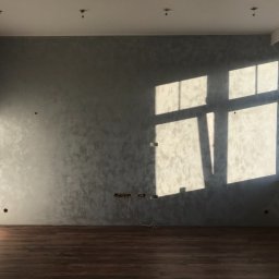 Krzysztof Dratwa - Pokój po gładziowaniu ścian w odcieniach szarości, z drewnianą podłogą i oknem, przez które wpada światło słoneczne. Widoczne puszki elektryczne.