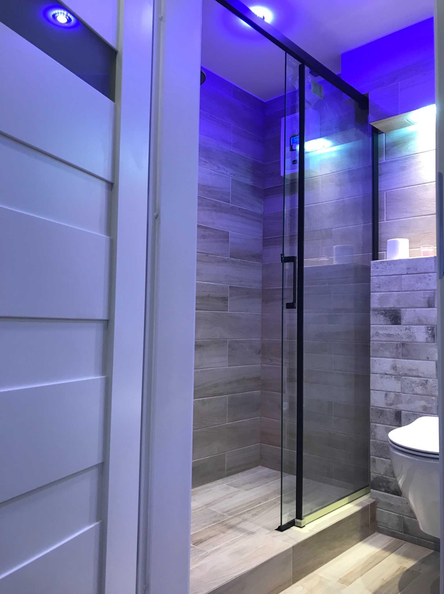 Nowoczesna łazienka z kabiną prysznicową typu walk-in, wykończona płytkami imitującymi drewno i oświetleniem LED w odcieniach niebieskiego. Widoczna toaleta podwieszana.