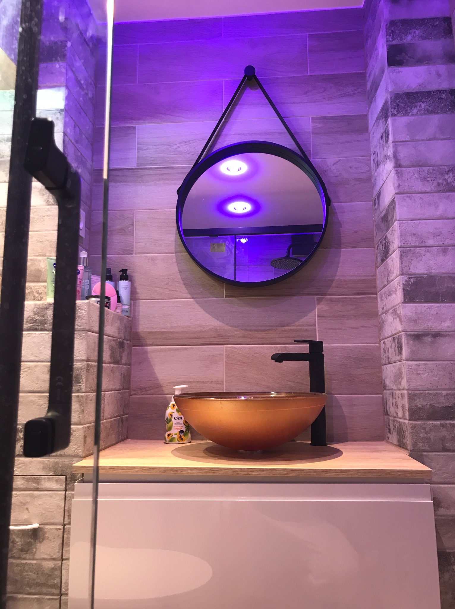 Nowoczesna łazienka z okrągłym lustrem na tle płytek imitujących cegłę, umywalką w odcieniach brązu i czarną armaturą, oświetlona fioletowym światłem.