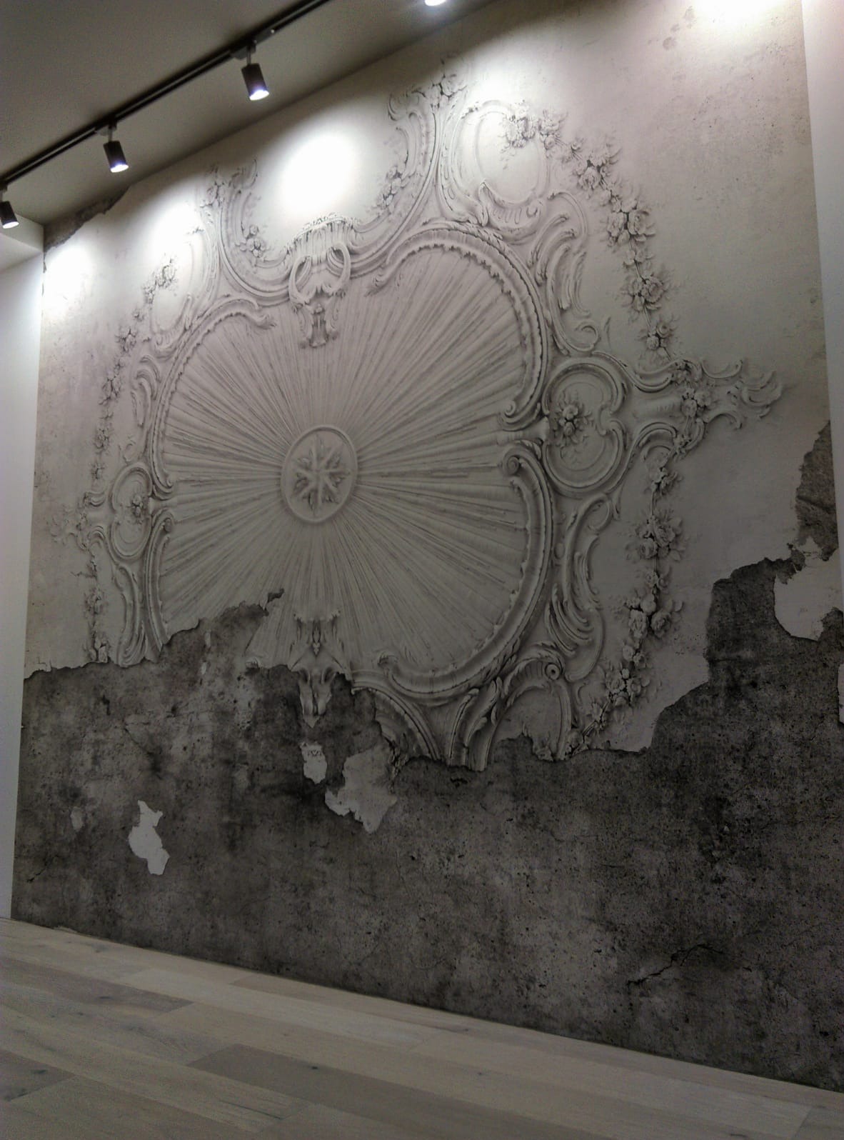Dekoracyjna ściana z efektem stiuku i przetarć, oświetlona punktowo. Ornament w stylu vintage z motywami roślinnymi. Widoczne fragmenty surowego tynku.