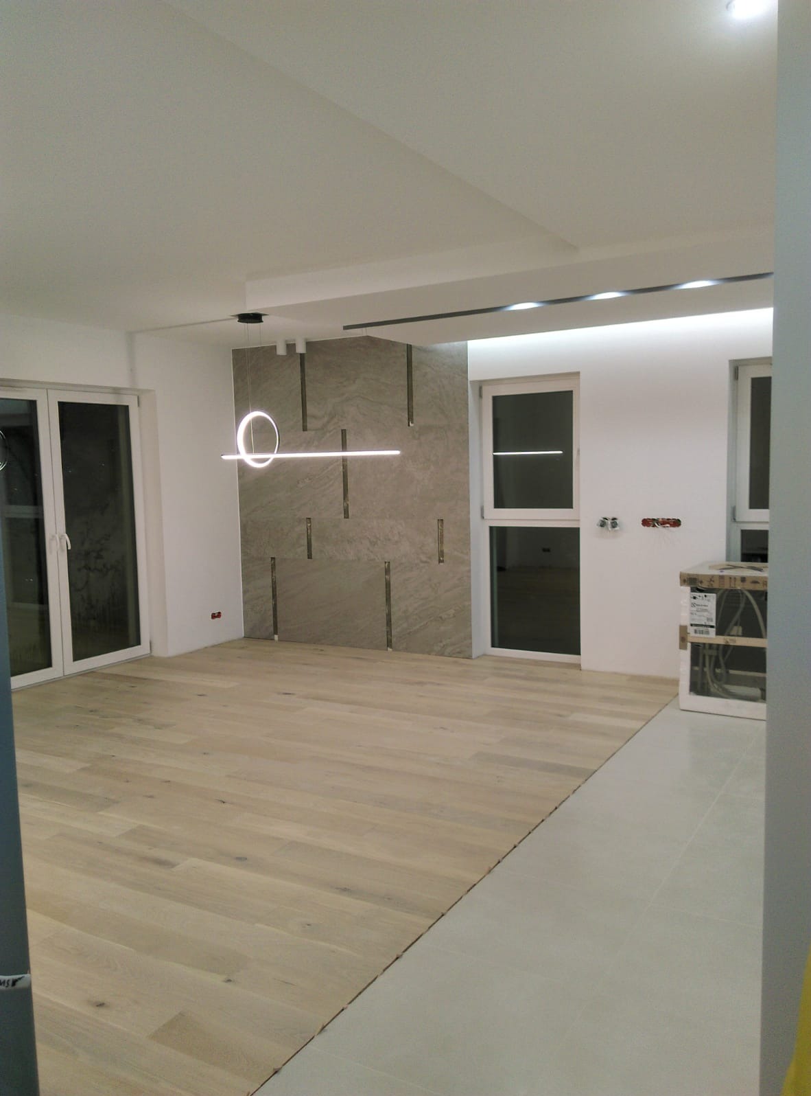 Jasne drewniane panele podłogowe w przestronnym pokoju z dekoracyjną ścianą z płytek i nowoczesnym oświetleniem. Widoczne okna i fragmenty instalacji elektrycznej.