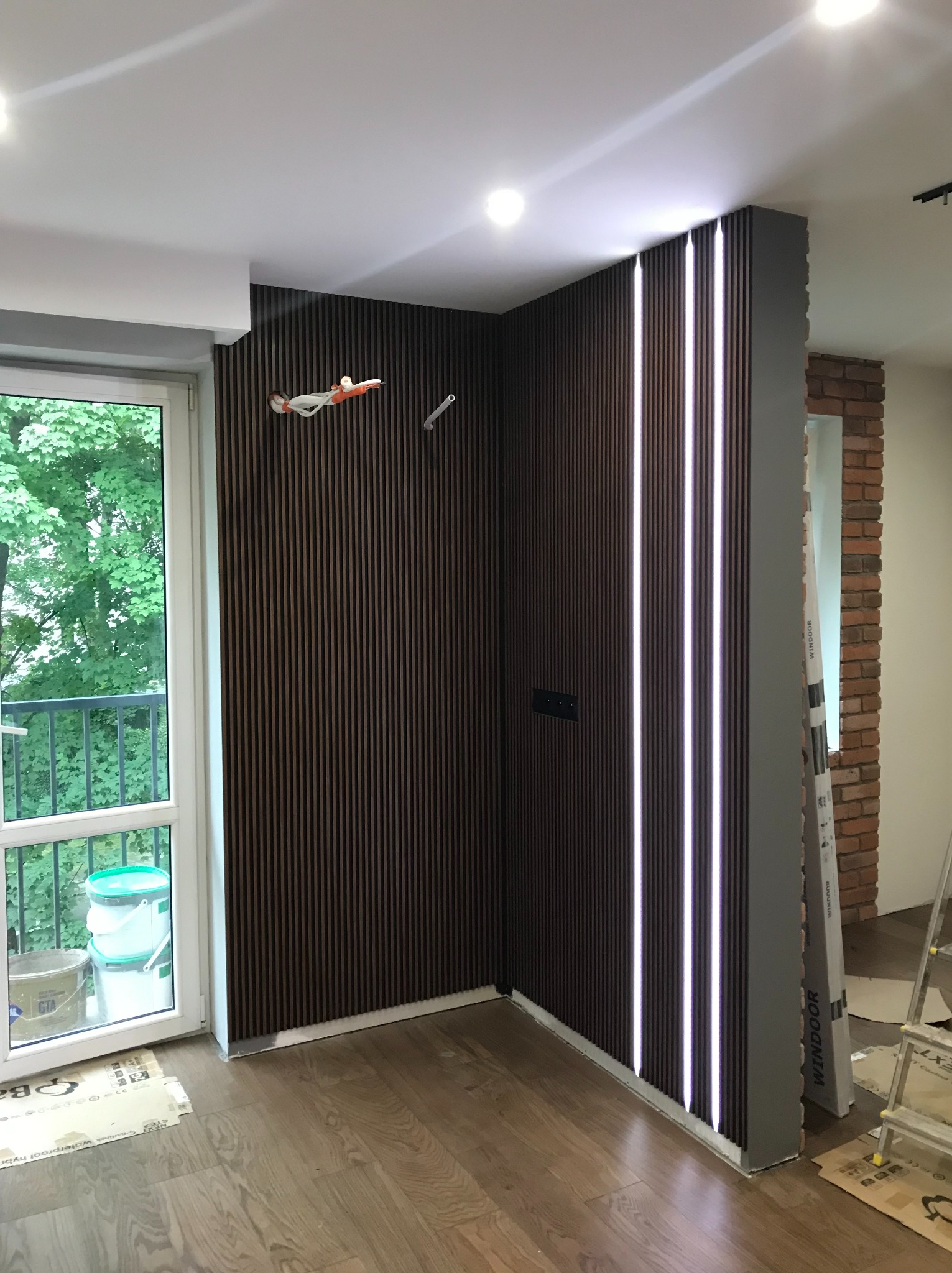 Nowoczesne wykończenie wnętrza z lamelami ściennymi w odcieniach brązu i oświetleniem LED. Widoczny fragment ceglanej ściany i okno balkonowe z widokiem na zieleń.