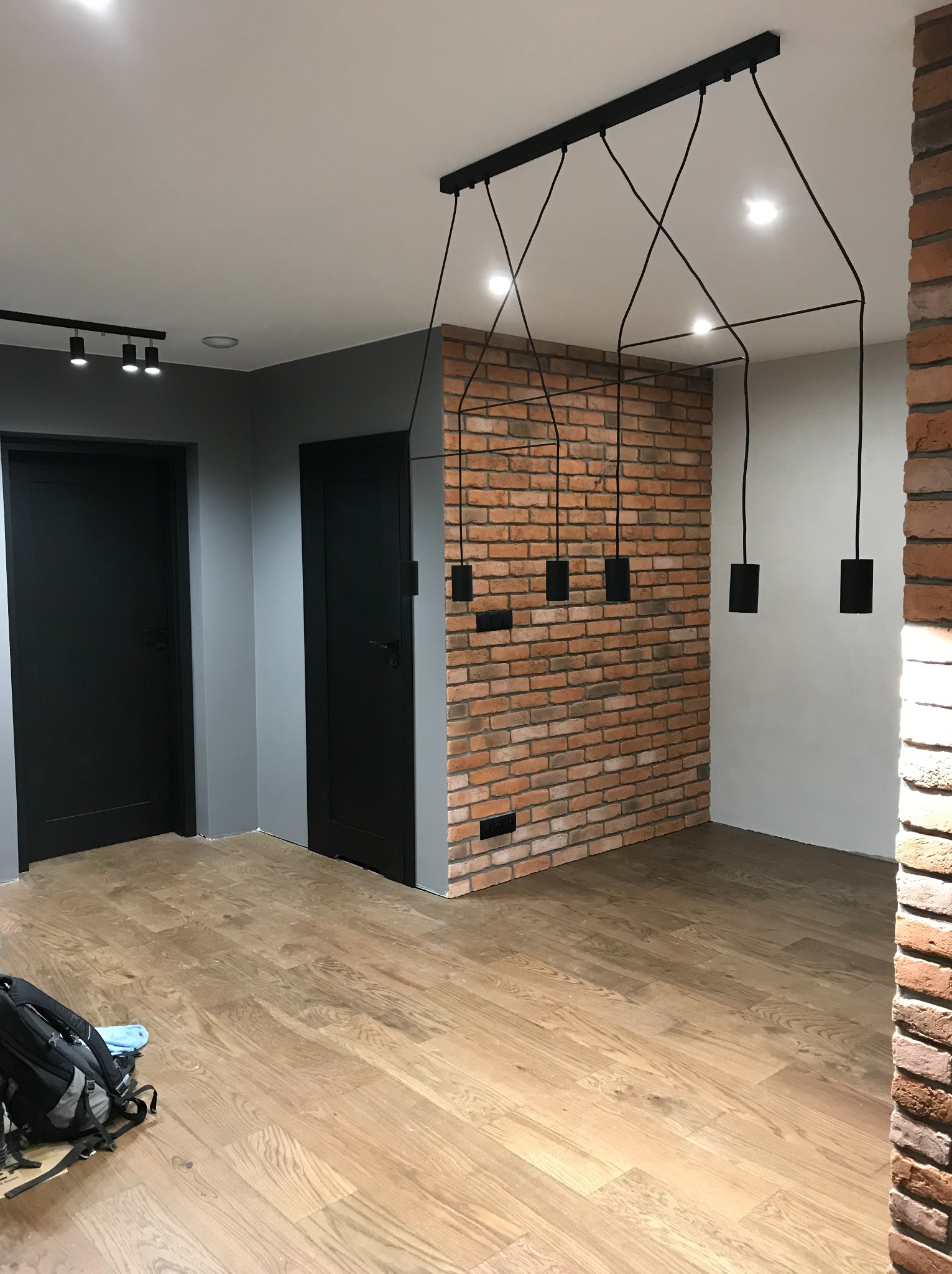 Nowoczesne wnętrze z ceglaną ścianą, wiszącą lampą z czarnymi kablami i drewnianą podłogą. Dwa czarne drzwi w szarej ścianie. Torba robocza w rogu.