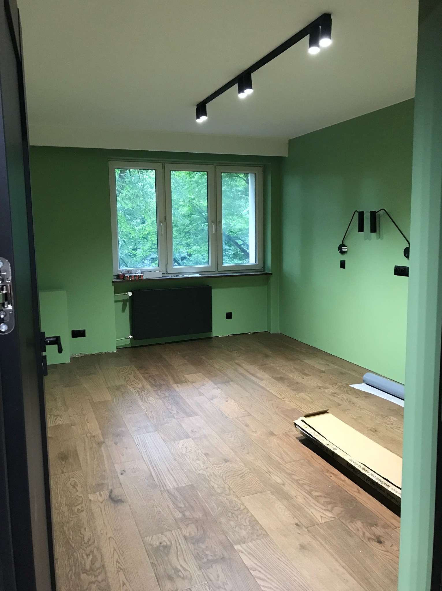 Pokój po odświeżeniu: zielone ściany, drewniana podłoga, nowoczesne oświetlenie. Widoczne listwy przypodłogowe i grzejnik. Częściowo widoczna kartonowa paczka z panelami.