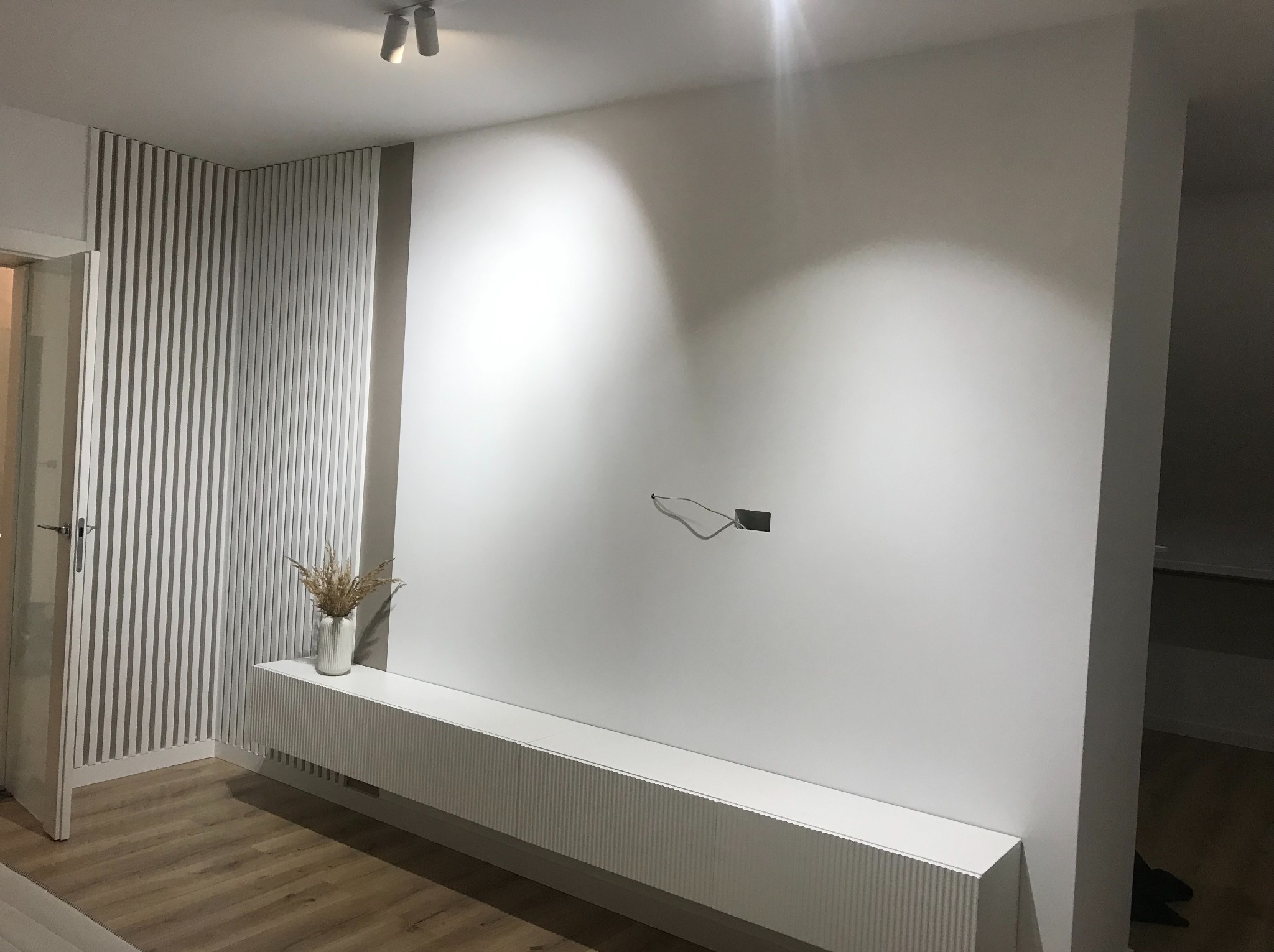 Biała ściana z listwami dekoracyjnymi i szafką RTV w Będzinie. Minimalistyczny design, drewniana podłoga, nowoczesne oświetlenie punktowe. Widoczne kable.