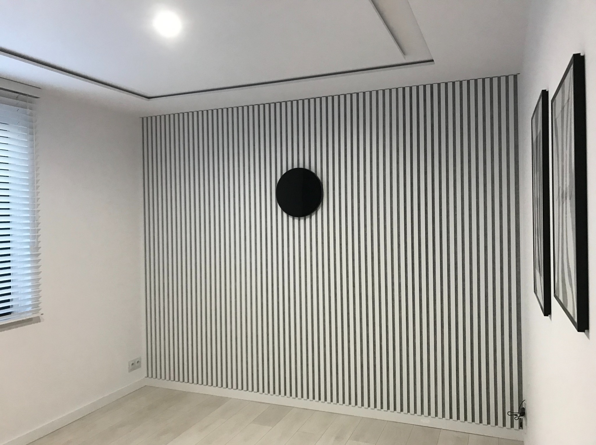 Nowoczesna ściana z pionowych paneli dekoracyjnych w odcieniach szarości, z okrągłym, czarnym elementem centralnym i minimalistycznymi obrazami w czarnych ramkach.