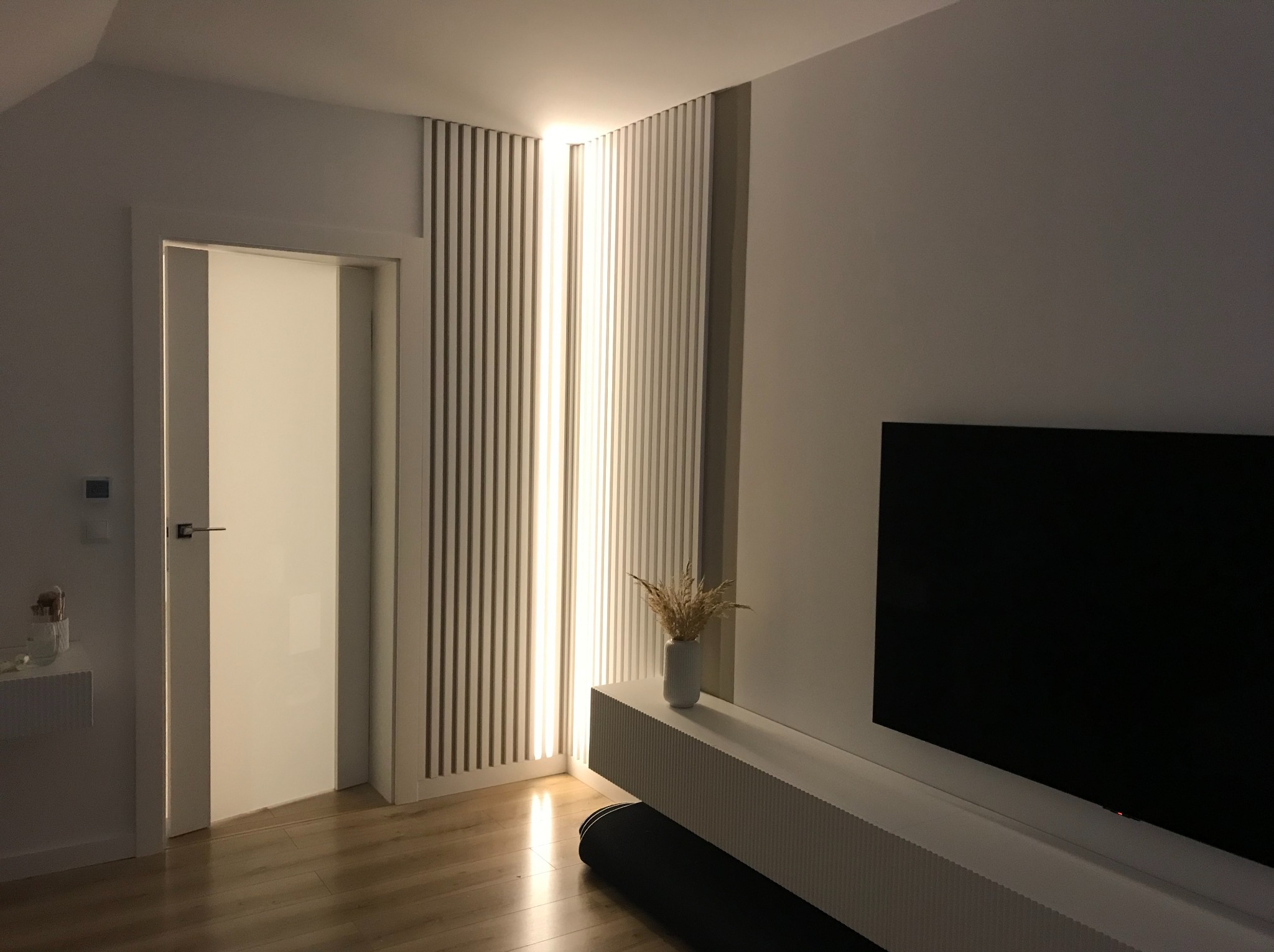 Nowoczesny salon z lamelami ściennymi podświetlonymi LED, białą szafką RTV i minimalistycznym wystrojem. Widoczna podłoga z paneli i fragment drzwi.