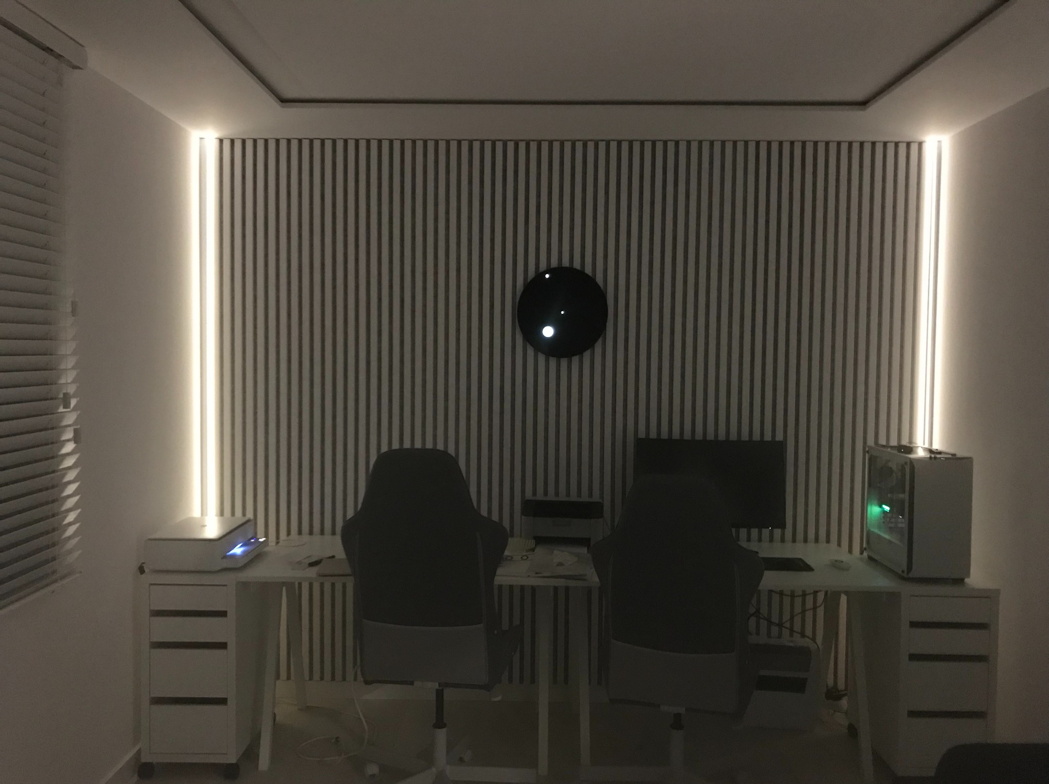 Pokój gracza z podświetlaną ścianą w pionowe pasy i oświetleniem LED po bokach. Dwa stanowiska komputerowe z krzesłami gamingowymi i czarnym zegarem na ścianie.