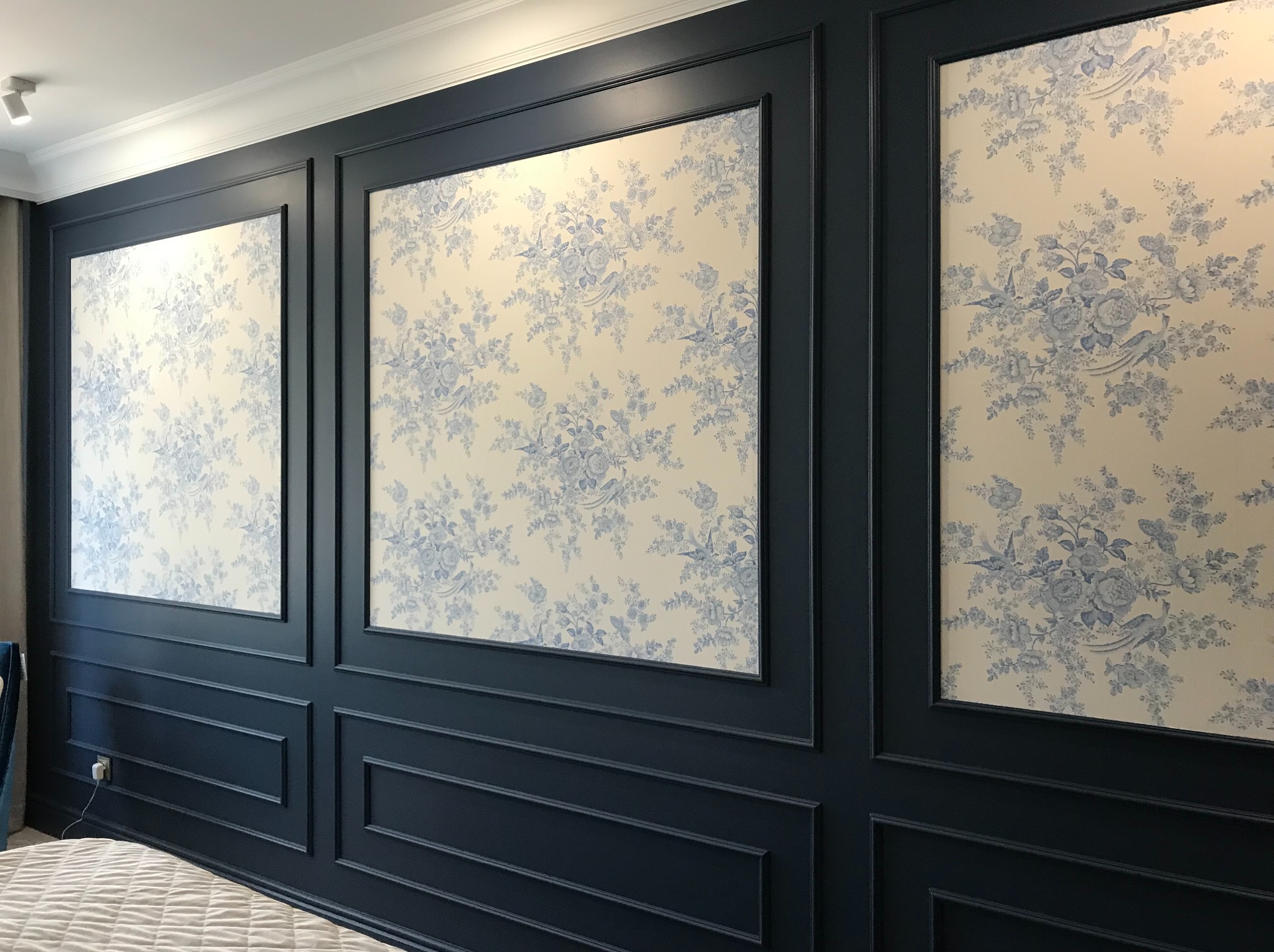 Elegancka ściana z ciemnoniebieskimi panelami i wstawkami z tapetą w delikatne, błękitne kwiaty. Klasyczny design wnętrza, subtelne oświetlenie.