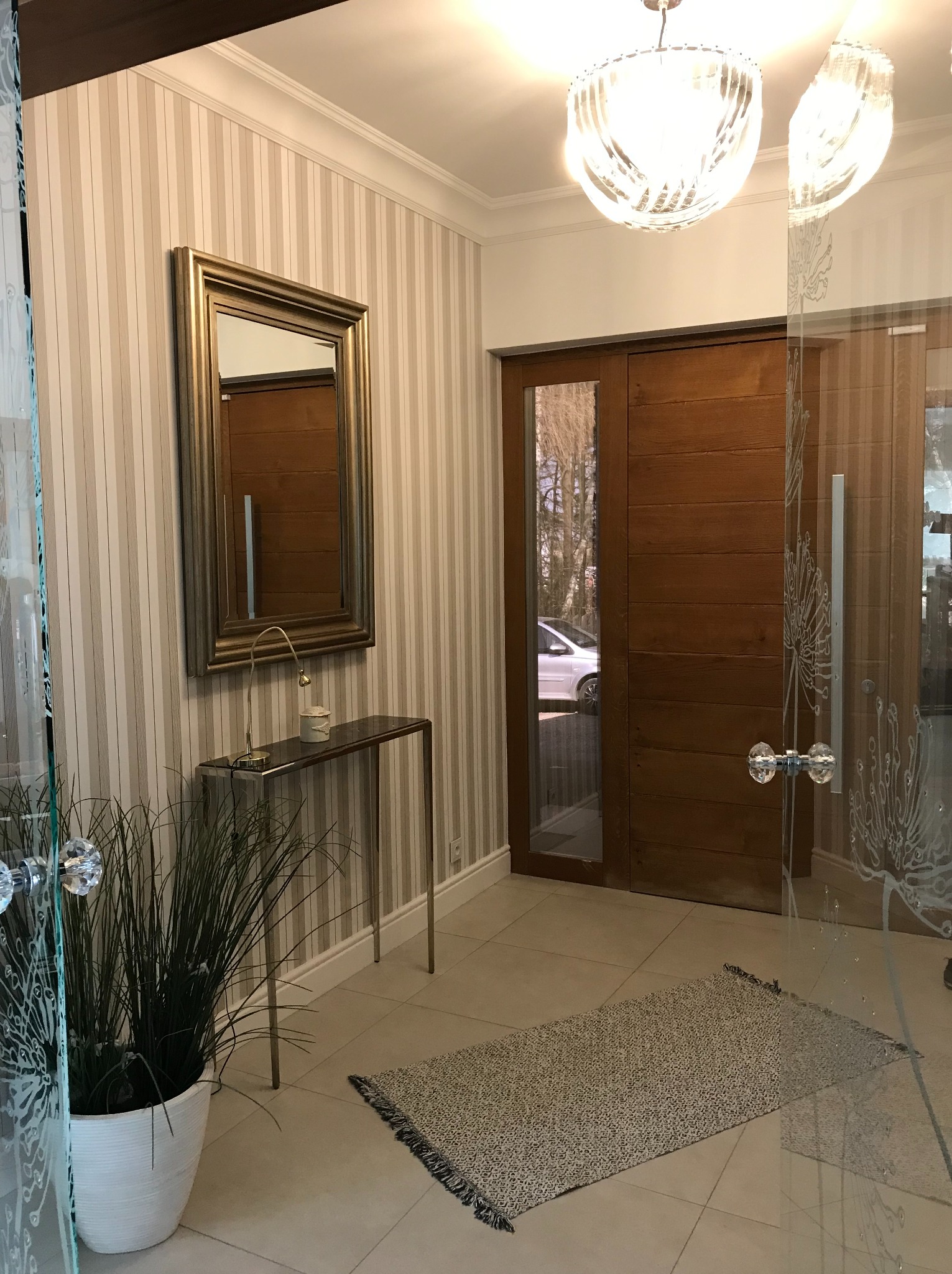 Elegancki hol z lustrem w złotej ramie, paskowaną tapetą i szklanymi drzwiami z dekoracyjnym motywem. Dwie lampy sufitowe oświetlają przestrzeń.