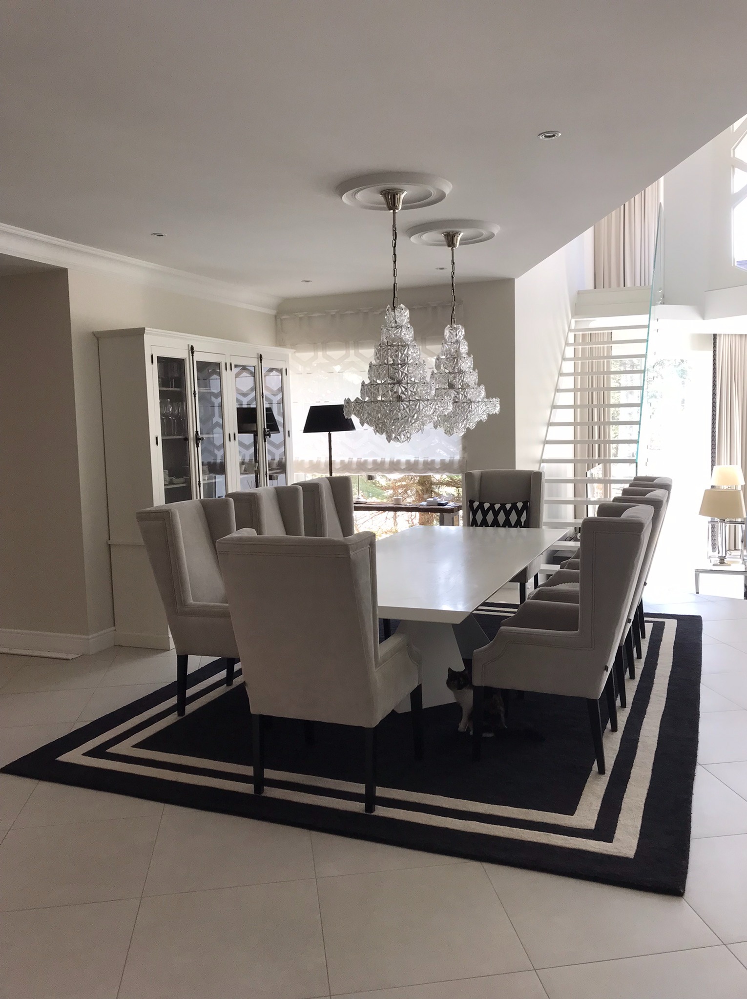 Jasne wnętrze jadalni z białym stołem, krzesłami w odcieniach szarości i kryształowymi żyrandolami. W tle białe schody i przeszklona witryna.