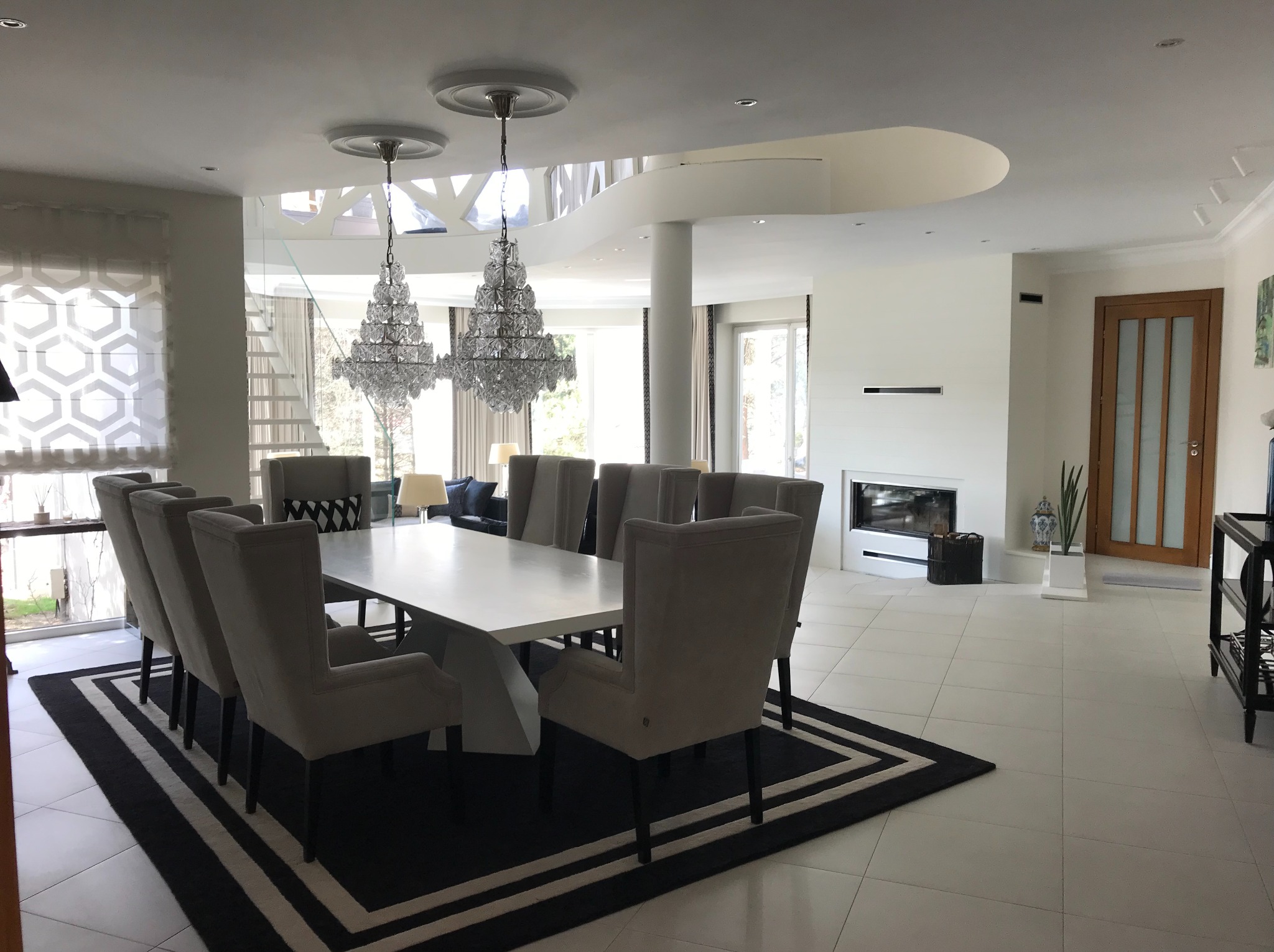 Jasne wnętrze jadalni z białymi ścianami, podłogą z płytek i dużym stołem otoczonym krzesłami. Dwa kryształowe żyrandole wiszą nad stołem. W tle kominek i schody.