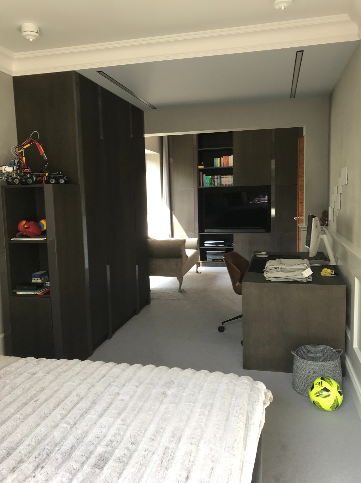 Nowoczesny pokój z zabudową meblową w odcieniach szarości i brązu. Widoczny fragment łóżka, biurko z komputerem, szafa i regał z książkami. Całość utrzymana w stonowanej kolorystyce.