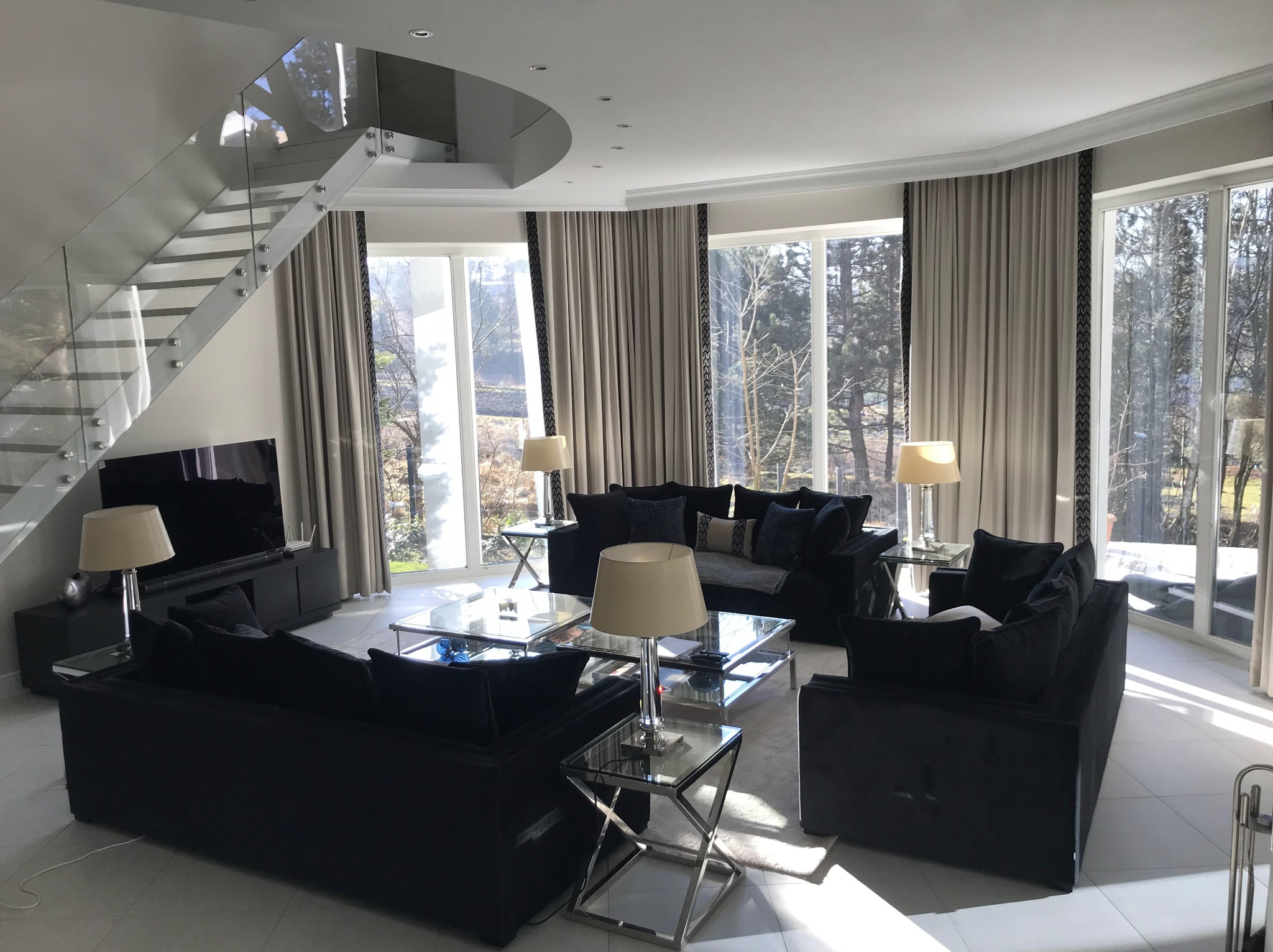 Elegancki salon z czarnymi sofami, szklanymi stolikami i schodami z przezroczystego szkła. Duże okna z zasłonami w odcieniach beżu wpuszczają naturalne światło.