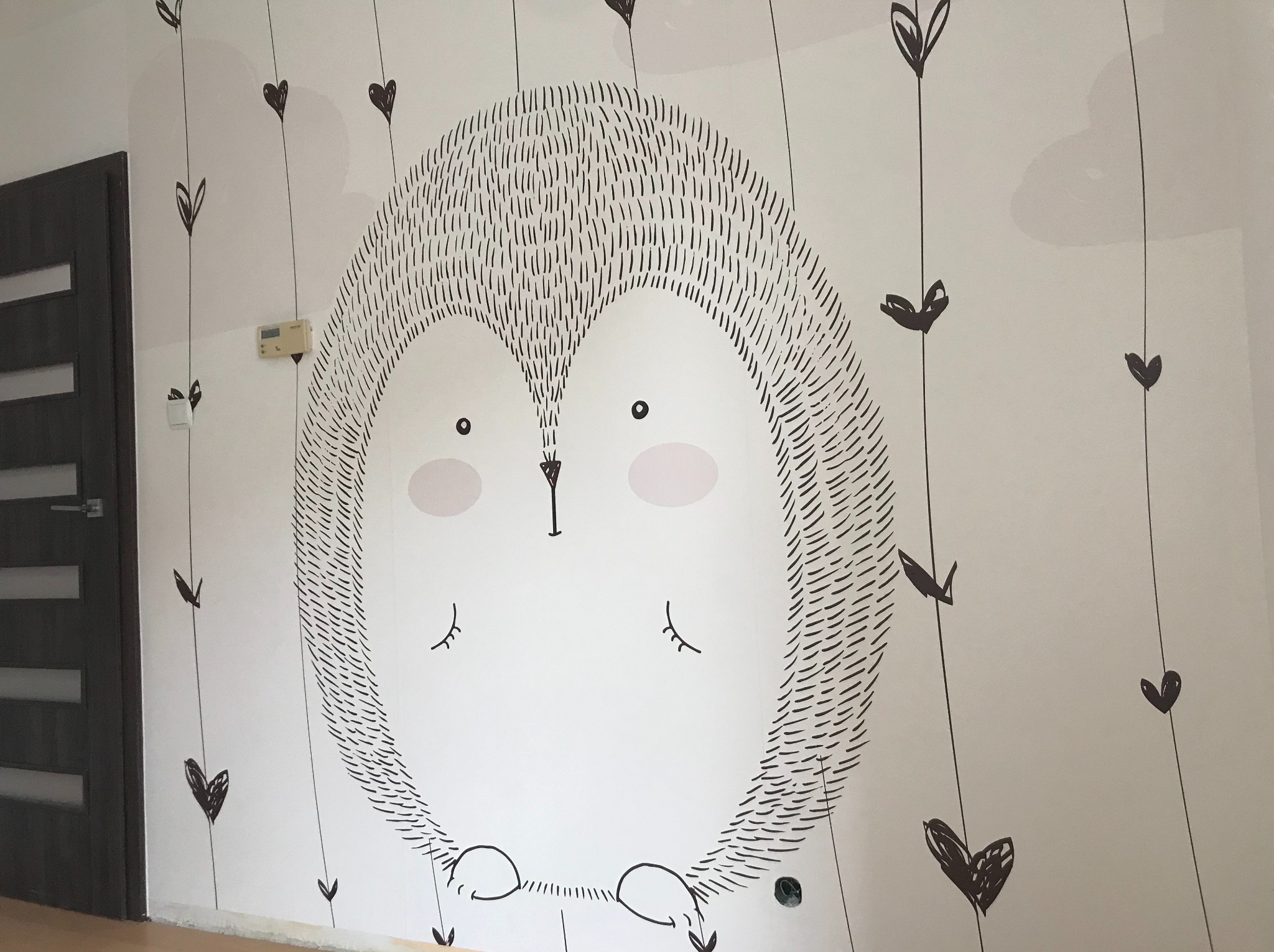 Ściana pokoju dziecięcego z ręcznie malowanym motywem jeża i serduszek. Minimalistyczny design, czarno-biała kolorystyka, widoczny fragment drzwi i podłogi.