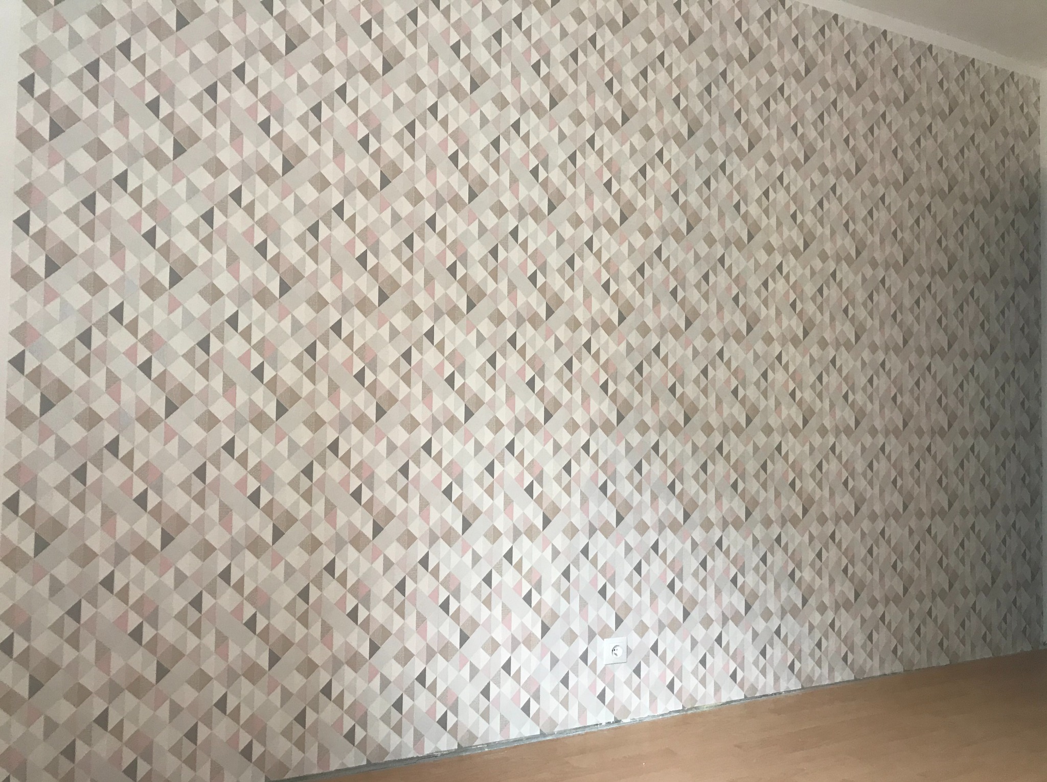 Ściana pokryta tapetą w geometryczny wzór z trójkątów w odcieniach szarości, beżu i różu. Widoczny fragment podłogi z paneli i gniazdko elektryczne.