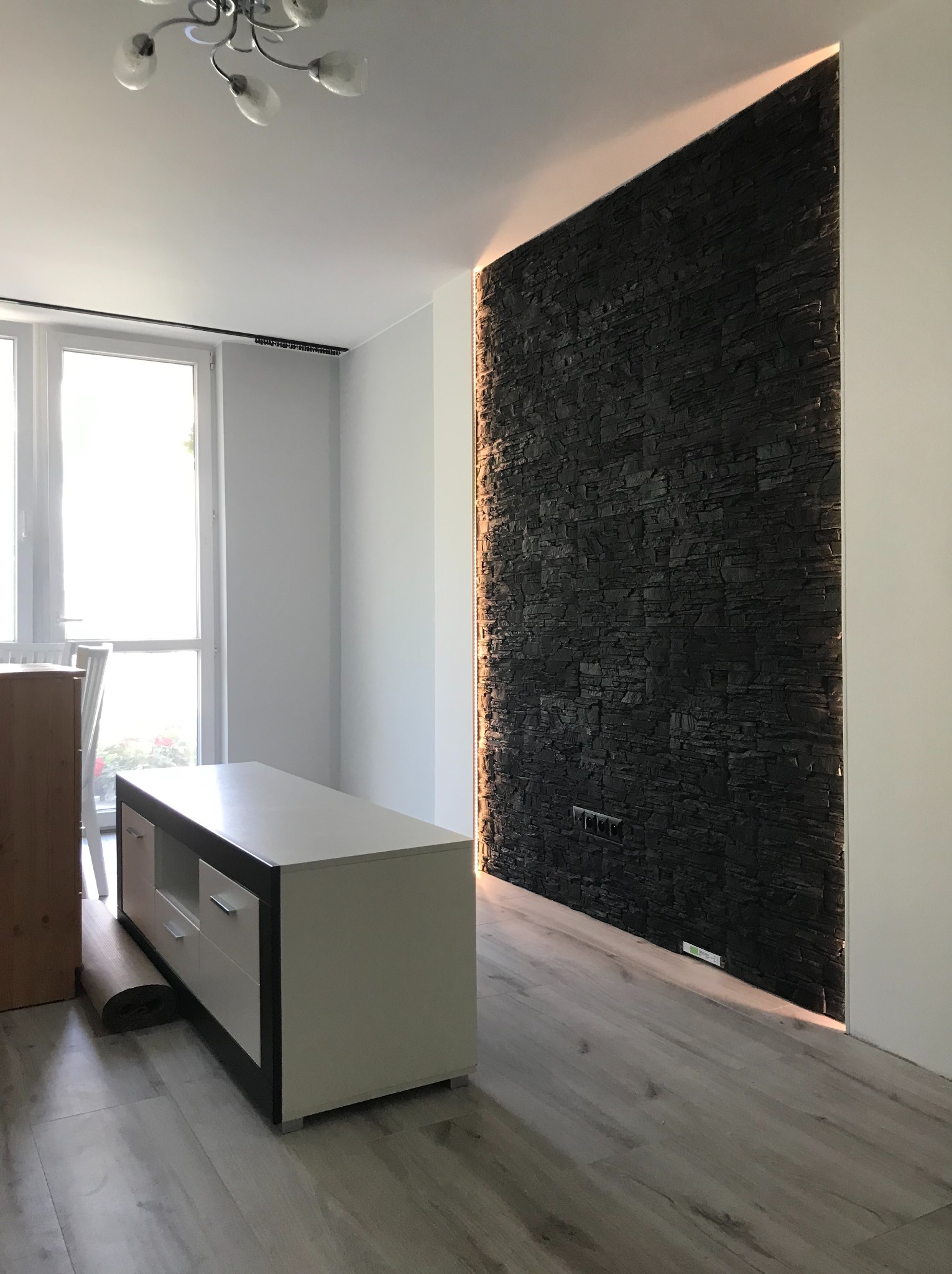 Nowoczesne wnętrze z akcentem: ściana wyłożona czarnym kamieniem dekoracyjnym, podświetlona po bokach, z widocznymi gniazdkami elektrycznymi i fragmentem mebla.