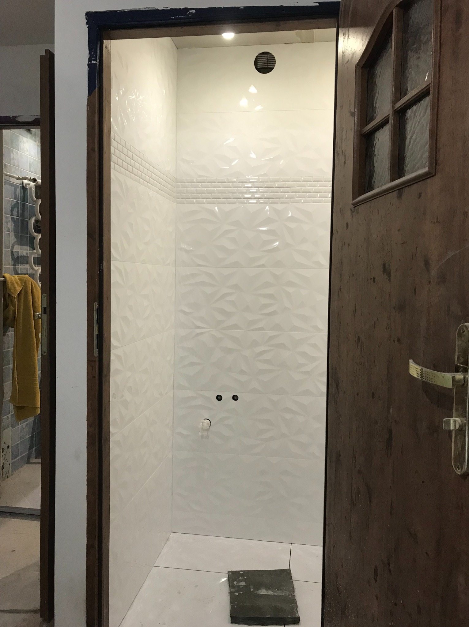 Wykończona kabina prysznicowa z białymi, trójwymiarowymi płytkami, widoczna przez otwarte, drewniane drzwi z szybą. W tle fragment innej łazienki.