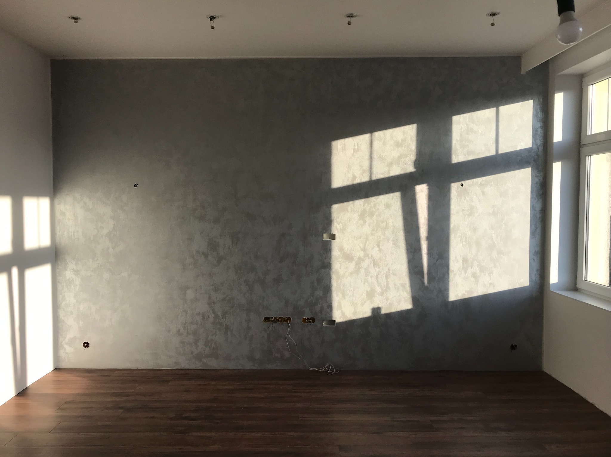 Pokój po gładziowaniu ścian w odcieniach szarości, z drewnianą podłogą i oknem, przez które wpada światło słoneczne. Widoczne puszki elektryczne.