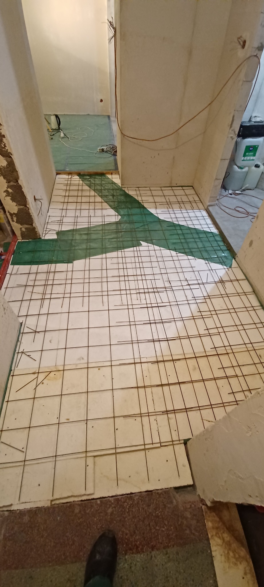 Przygotowanie podłoża pod wylewkę: białe płyty izolacyjne z siatką zbrojeniową, zielona folia. Widok z góry na surowy stan pomieszczenia w trakcie prac remontowych.