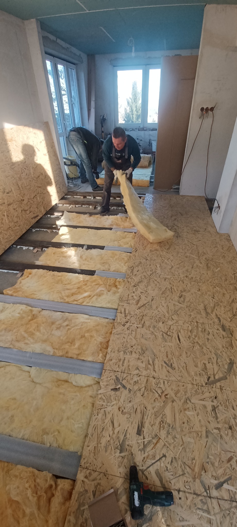 Ocieplanie podłogi wełną mineralną i płytami OSB. Dwóch monterów w trakcie pracy. Widoczne narzędzia i materiały budowlane. Jasne wnętrze w trakcie remontu.