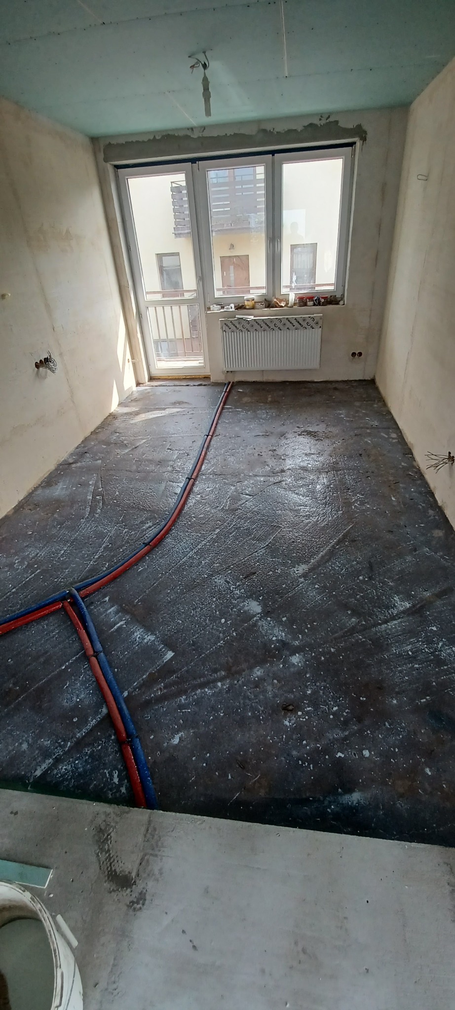 Wnętrze w trakcie remontu: widoczne rury ogrzewania podłogowego w wylewce, surowy tynk na ścianach i okno z widokiem na budynek. Etap prac przed wykończeniem.