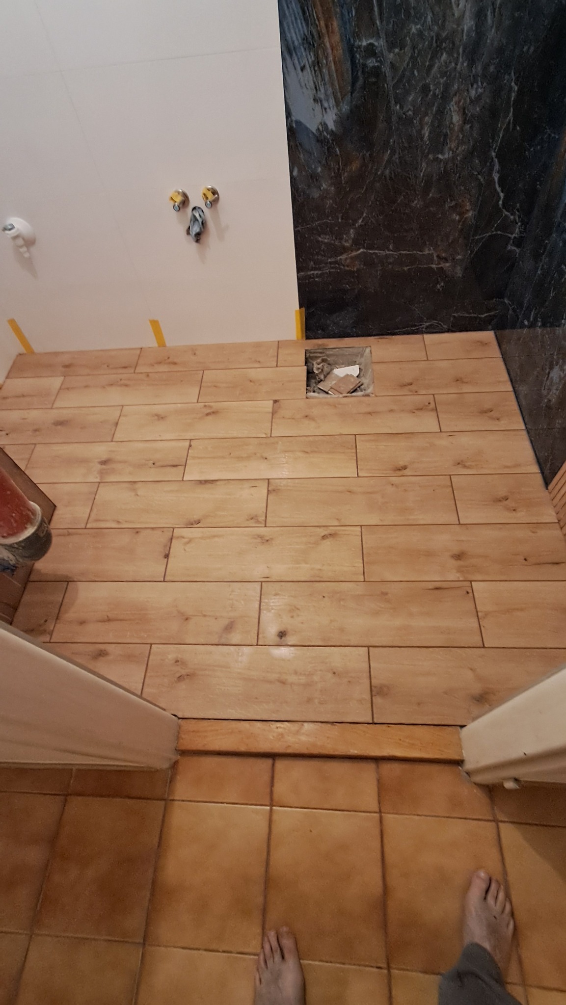 Ułożona podłoga z płytek imitujących drewno w łazience, widoczne fragmenty ściany z instalacją i otwór w podłodze. W tle ciemna ściana z wzorem marmuru.
