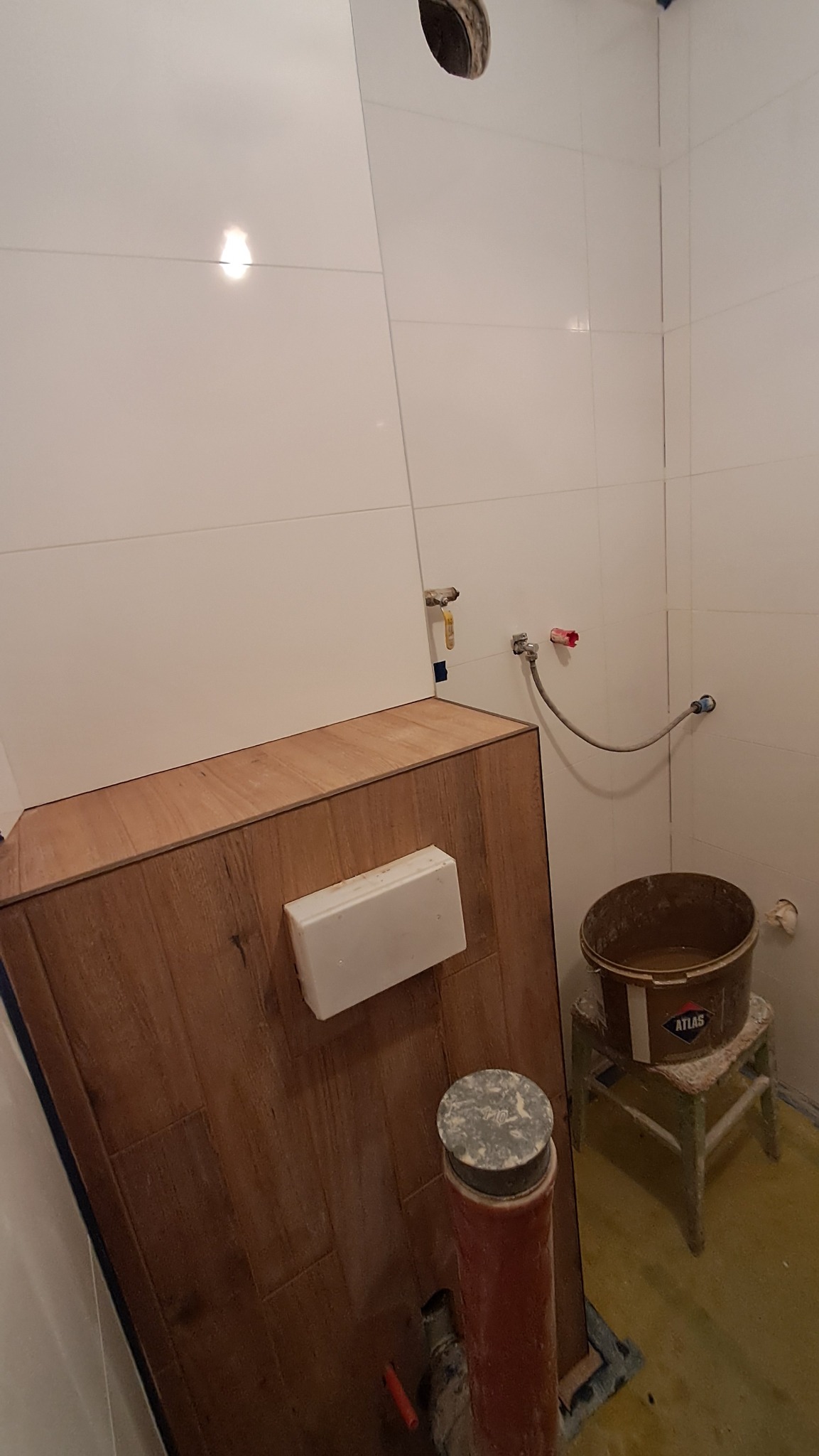 Białe płytki w łazience z drewnopodobną zabudową stelaża WC, widoczne rury i wiadro z zaprawą na stołku. Etap prac wykończeniowych.