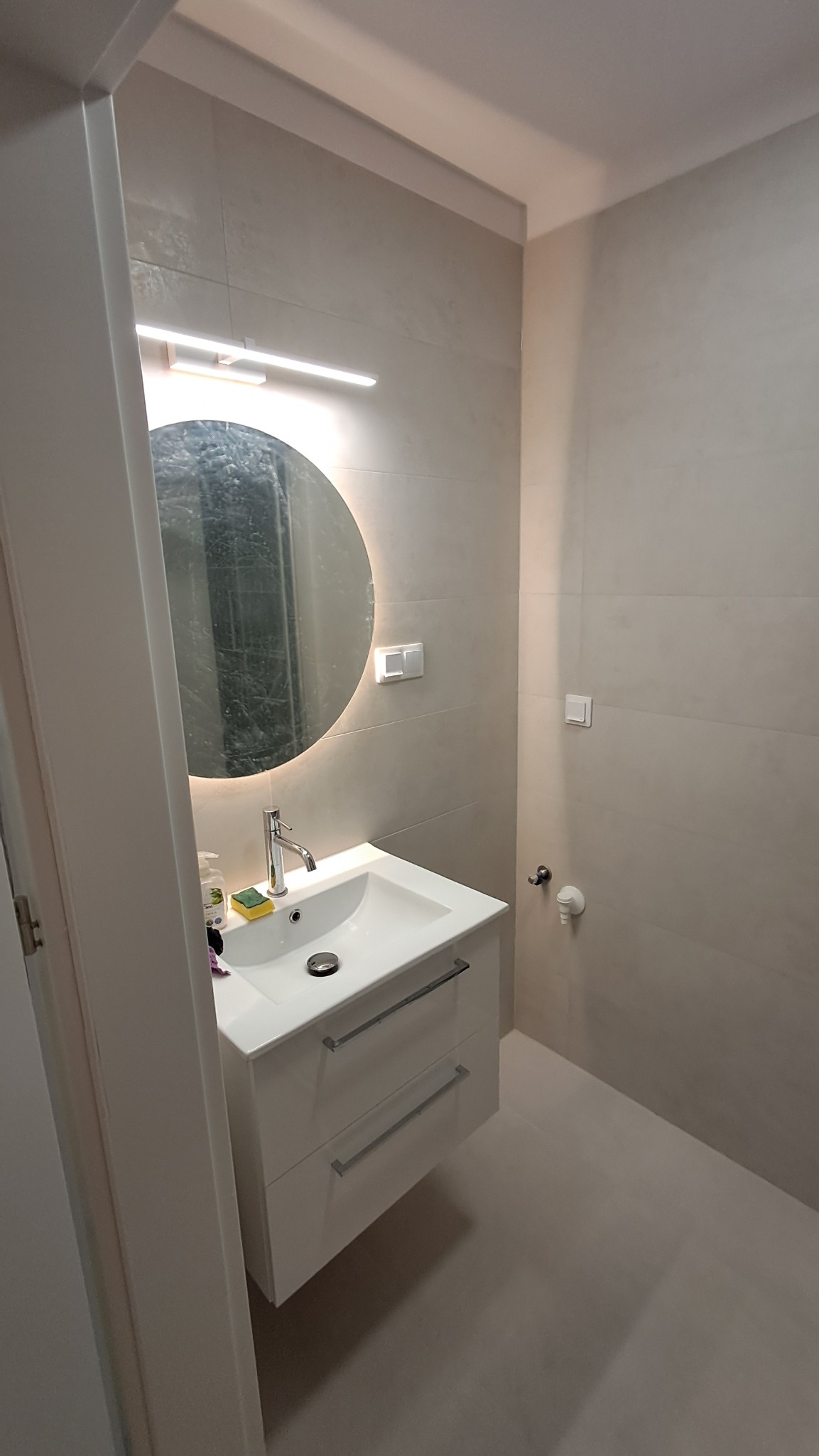 Nowoczesna łazienka w Wieliczce z okrągłym lustrem LED, białą szafką pod umywalką i beżowymi płytkami. Minimalistyczny design i subtelne oświetlenie.