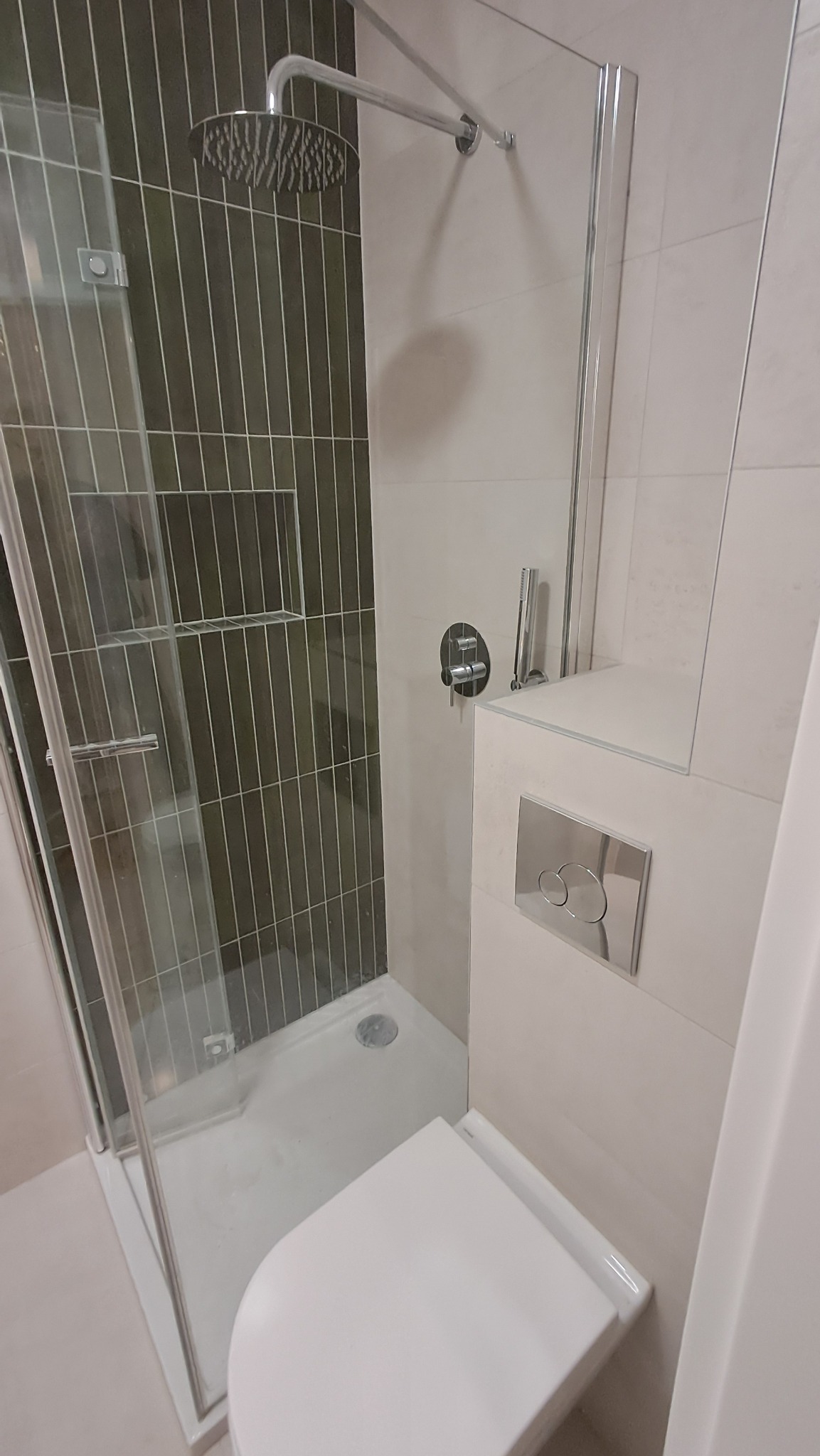 Nowoczesna kabina prysznicowa z deszczownicą i toaleta w wykończonej łazience. Ciemne, pionowe płytki w kabinie kontrastują z jasnymi płytkami na pozostałej części ściany.