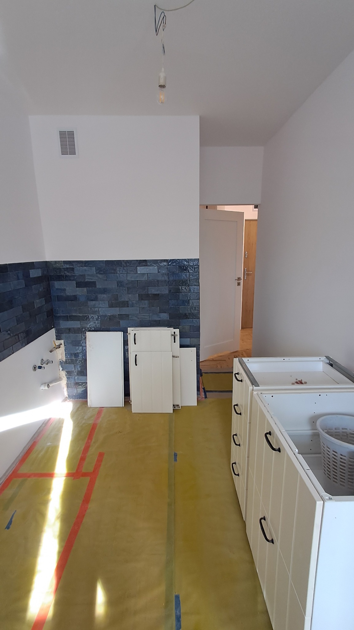 Wnętrze kuchni w trakcie remontu: ułożona glazura na ścianie, zabezpieczona podłoga folią, białe fronty szafek i niedokończona zabudowa. Widoczny surowy stan pomieszczenia.