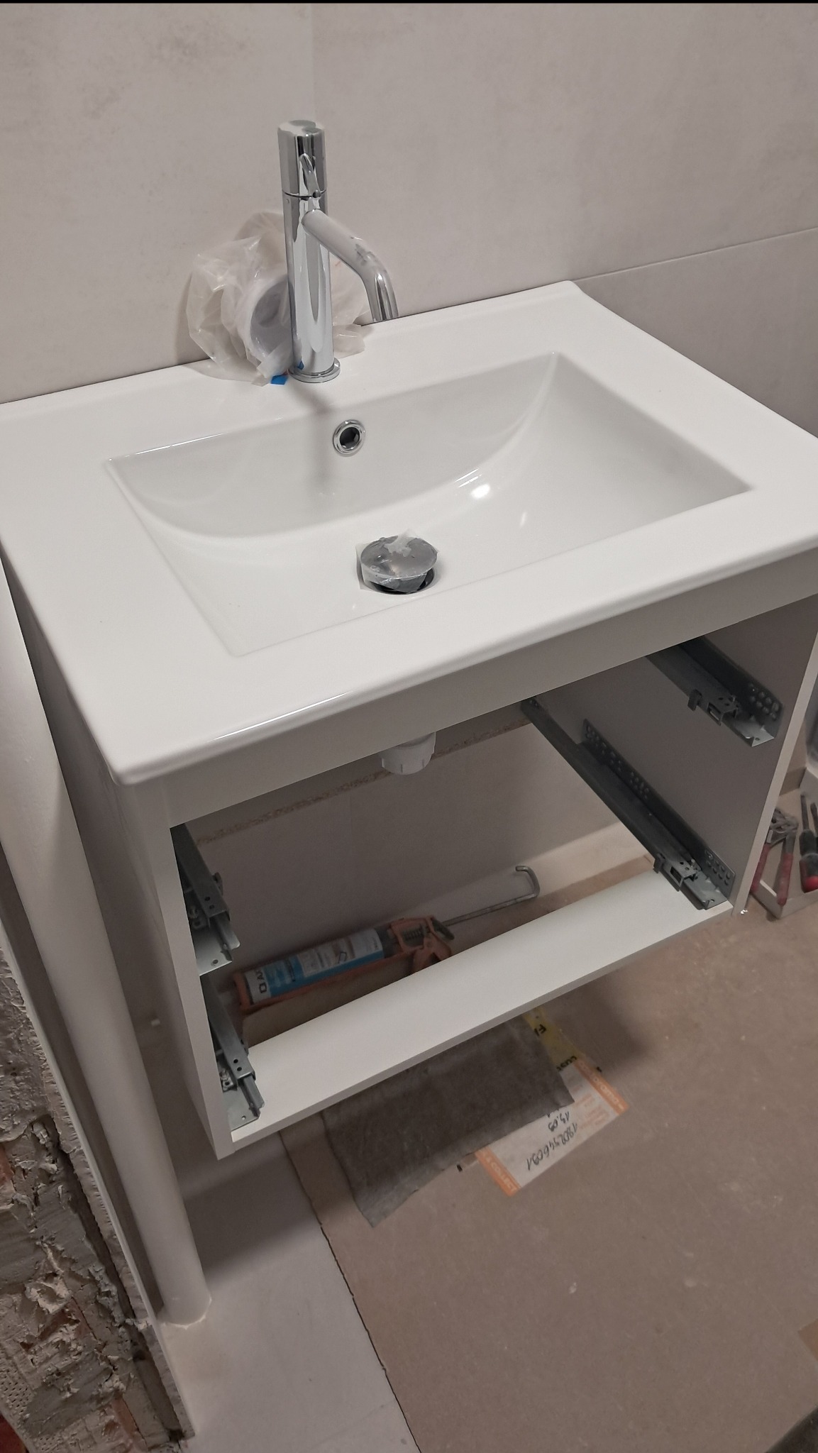 Biała umywalka z szafką w trakcie montażu, widoczne prowadnice szuflad i silikon. Świecąca bateria z folią ochronną. Fragment ściany z resztkami starej glazury.