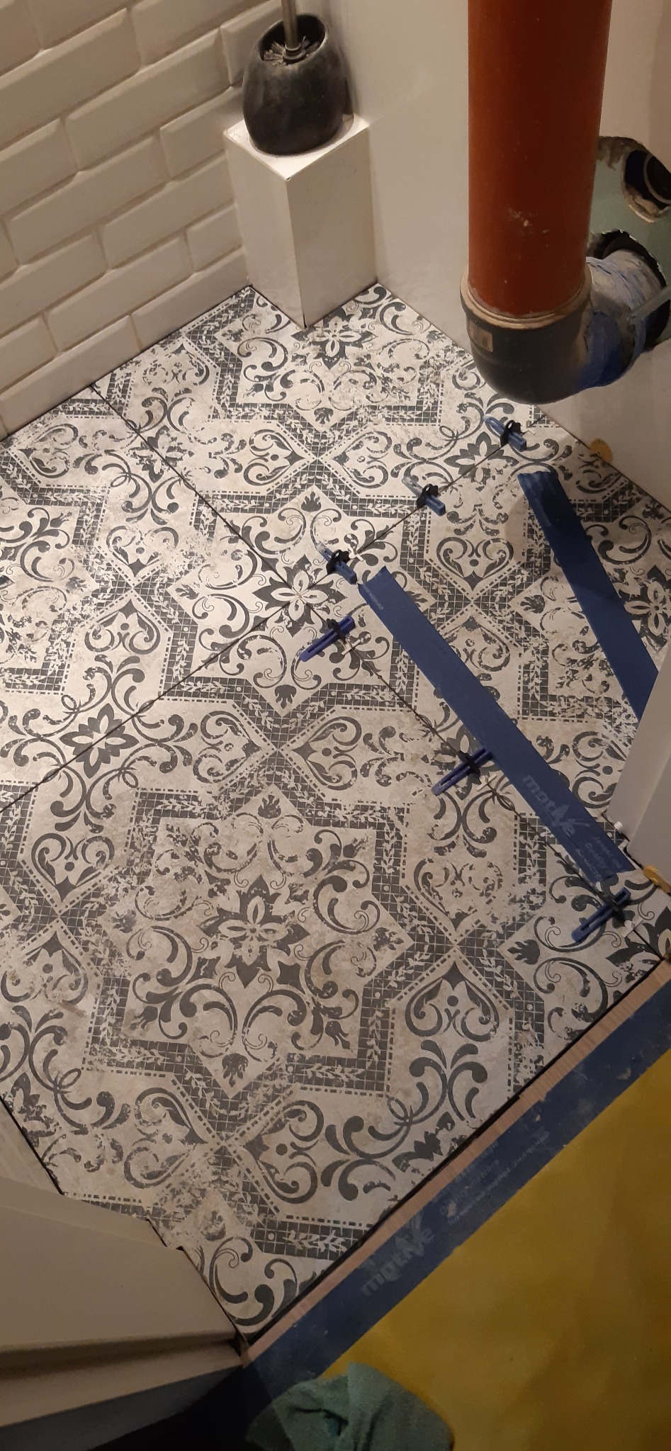 Układanie wzorzystych płytek podłogowych w łazience, z użyciem niebieskich klinów poziomujących. Widoczny fragment ściany z białej cegiełki i rura kanalizacyjna.
