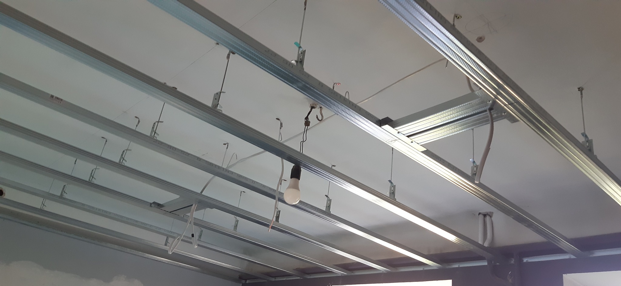 Metalowa konstrukcja sufitu podwieszanego z widocznymi profilami, przewodami i żarówką. Elementy montażowe w zbliżeniu, sufit w trakcie instalacji.