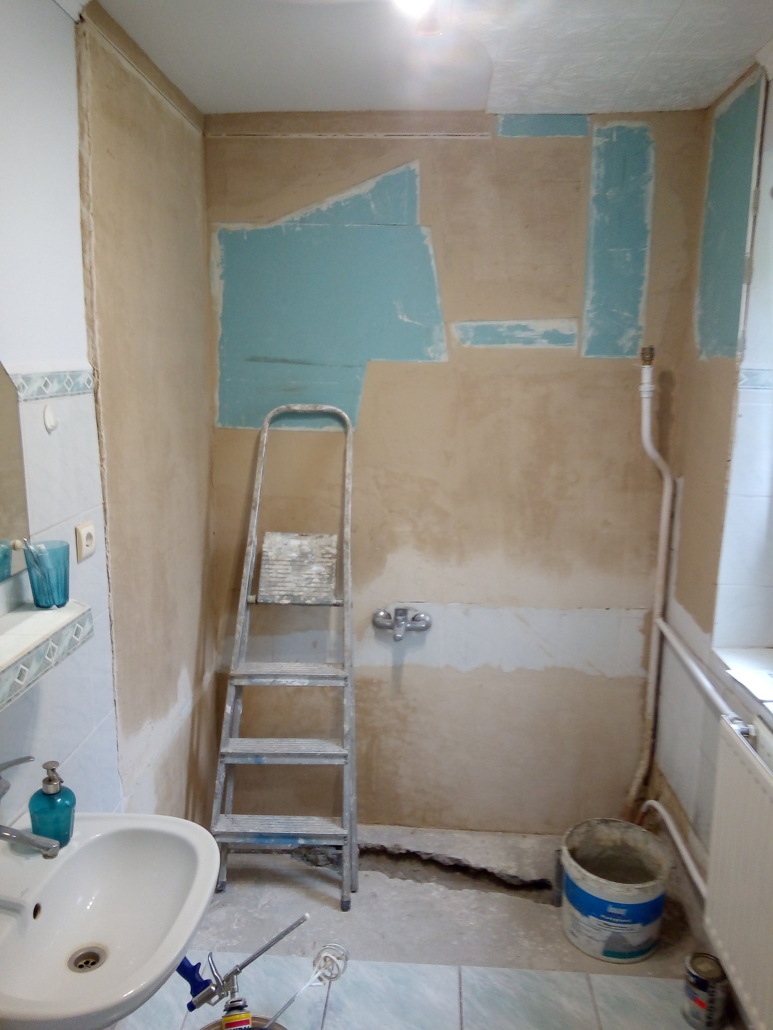 Łazienka w trakcie prac: ściany z fragmentami gładzi i niebieskiej farby, drabina, umywalka, narzędzia. Widoczny etap przygotowania do dalszych prac wykończeniowych.